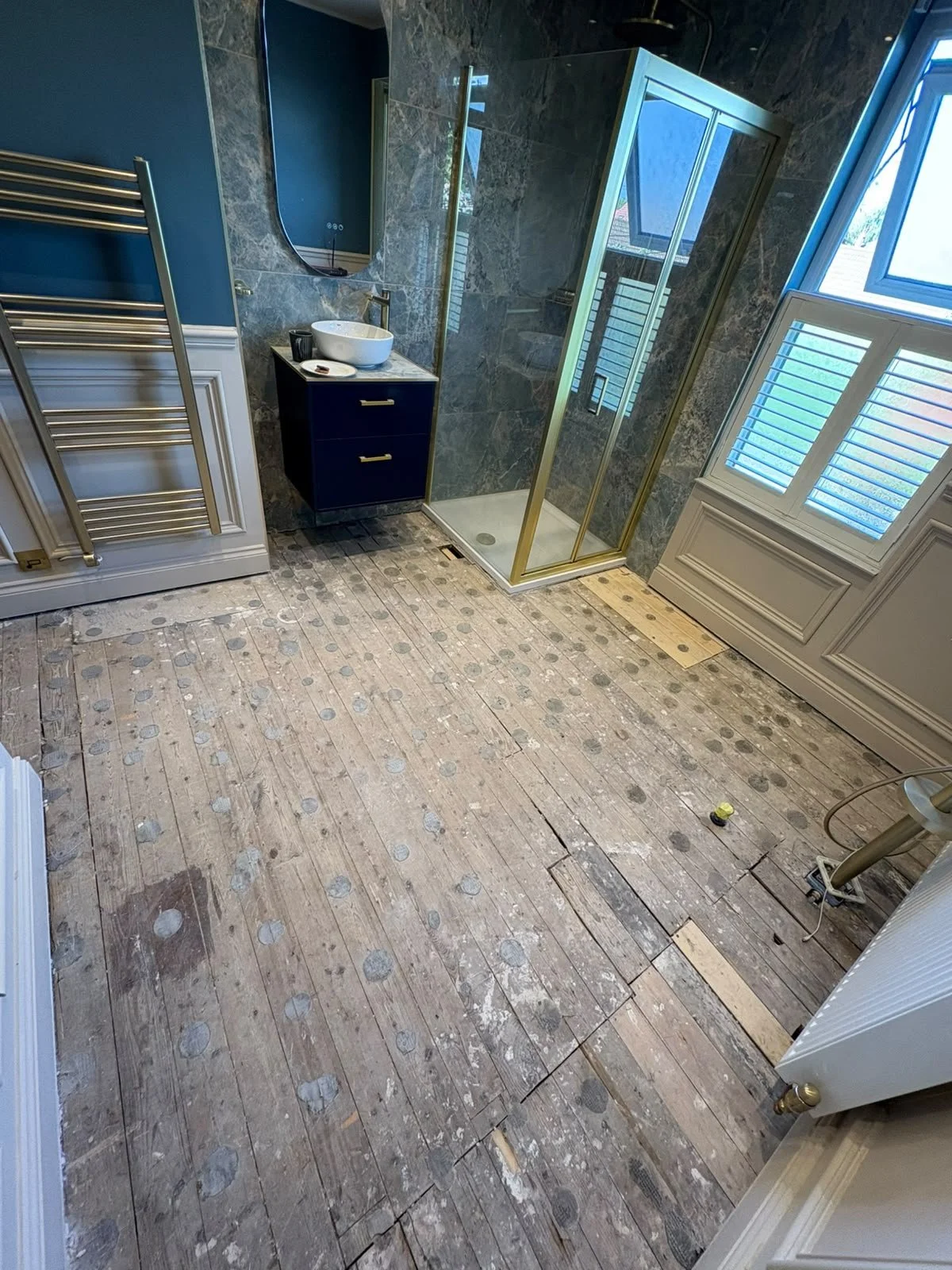 A bathroom under renovation with a wood floor, a small black vanity with a vessel sink, a mirror, a glass shower enclosure, a window with shutters, and a white radiator. The floor appears unfinished with visible floorboards and some construction debr