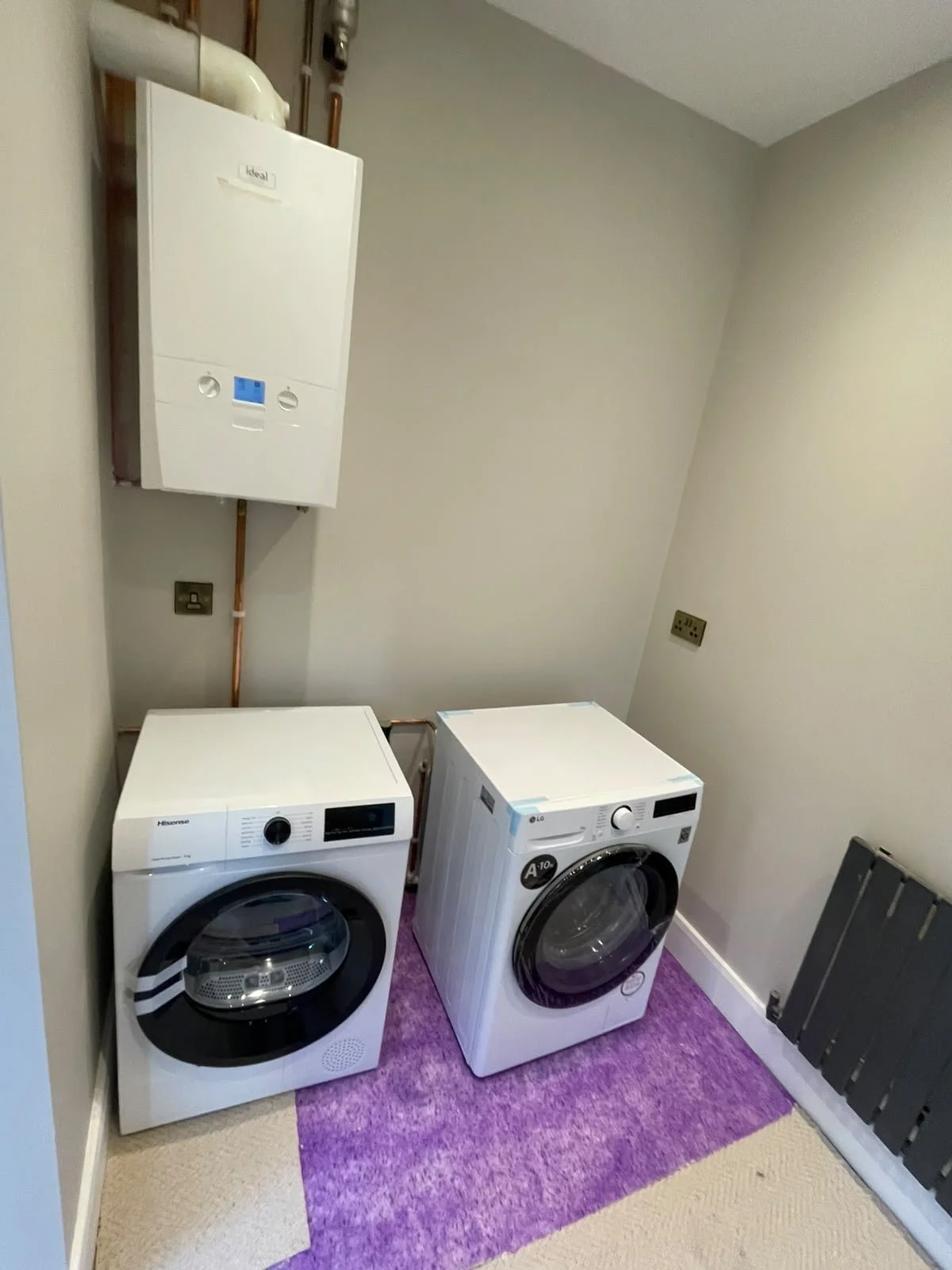 A laundry room with a washing machine and dryer on a purple rug, a wall-mounted boiler above, and electrical outlets on the wall.