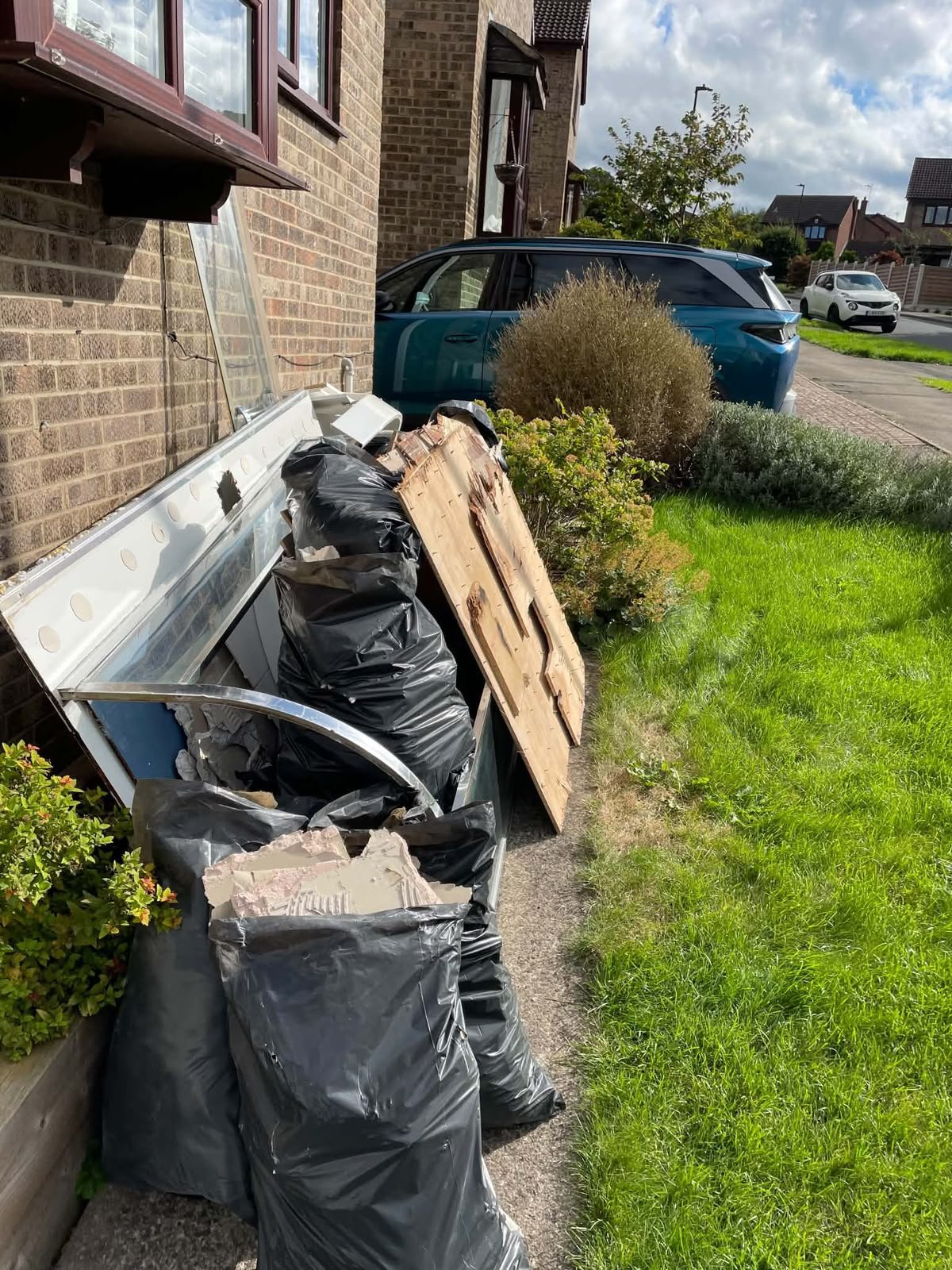 Trash bags, a piece of furniture, and a wooden board dumped along the side of a house with a garden, parked cars, and residential houses in the background.