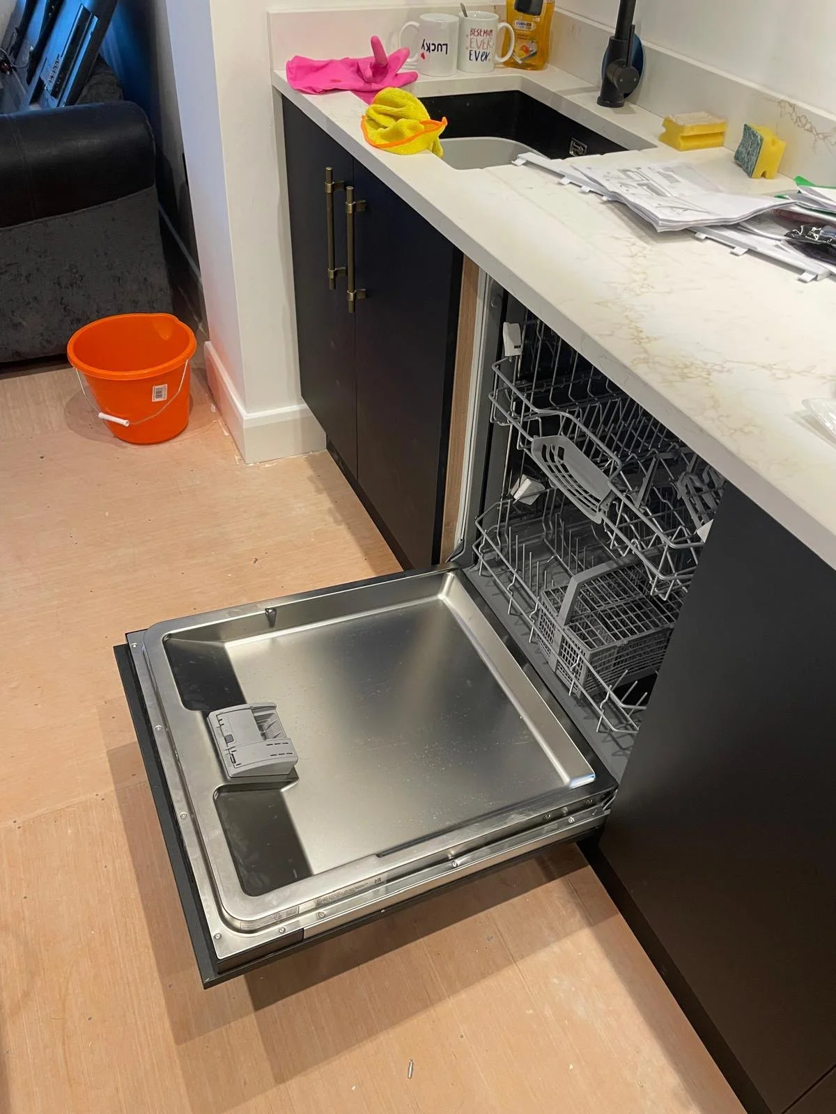 Empty dishwasher pulled out from under kitchen counter with open doors, near a black cabinet with items on the countertop including dish soap, paper towels, and mugs.