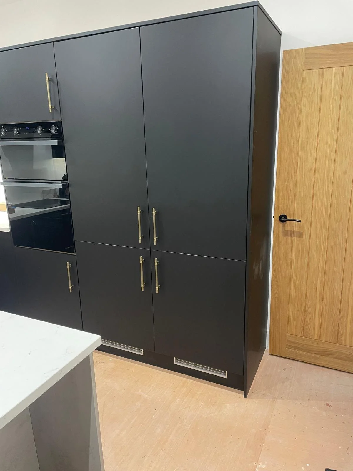 Black built-in kitchen cabinet with gold handles next to a wooden door with a black handle.