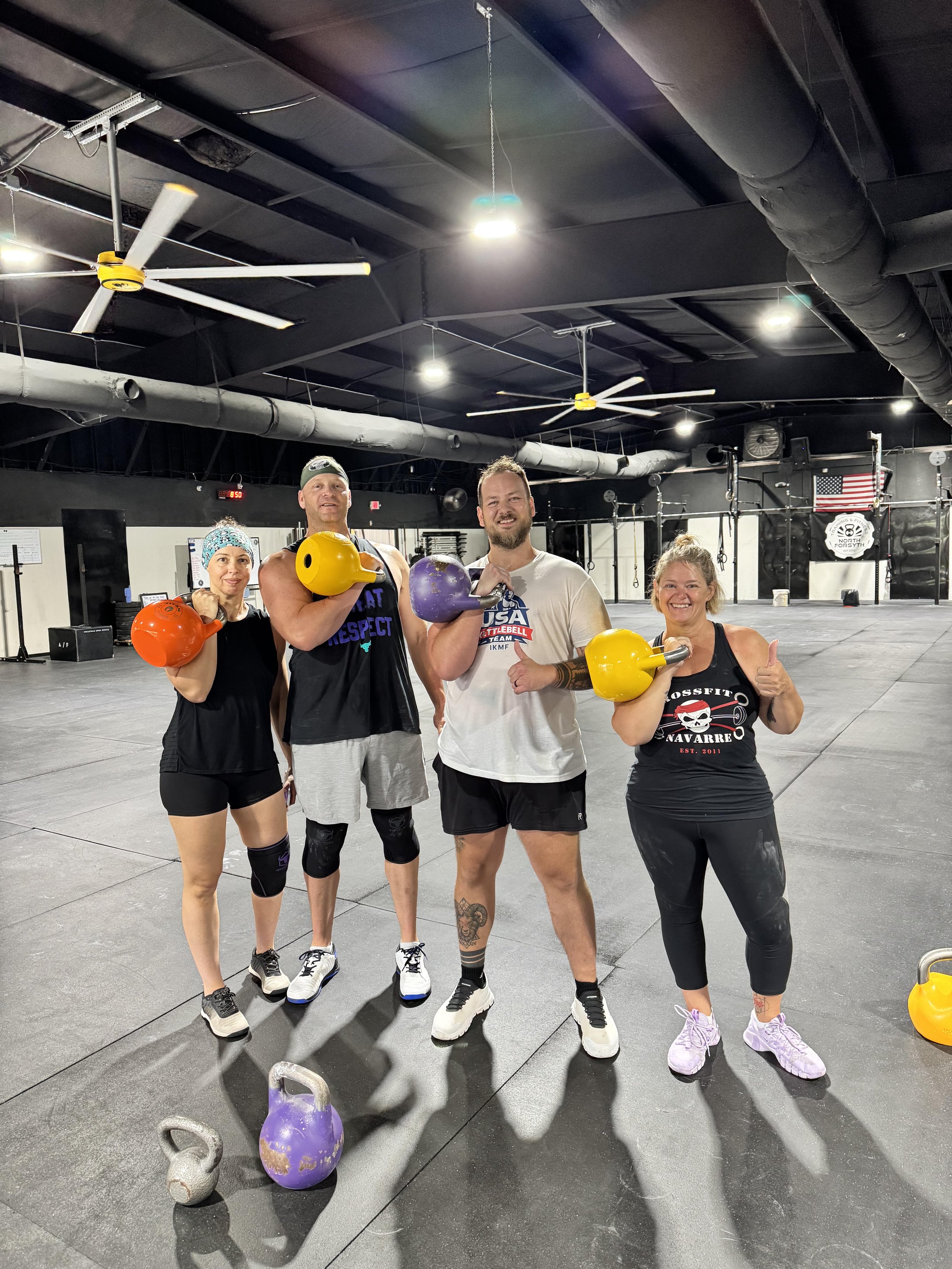 Four people stand in a gym holding kettlebells, smiling at the camera. The gym has black flooring, white walls, and large ceiling fans.