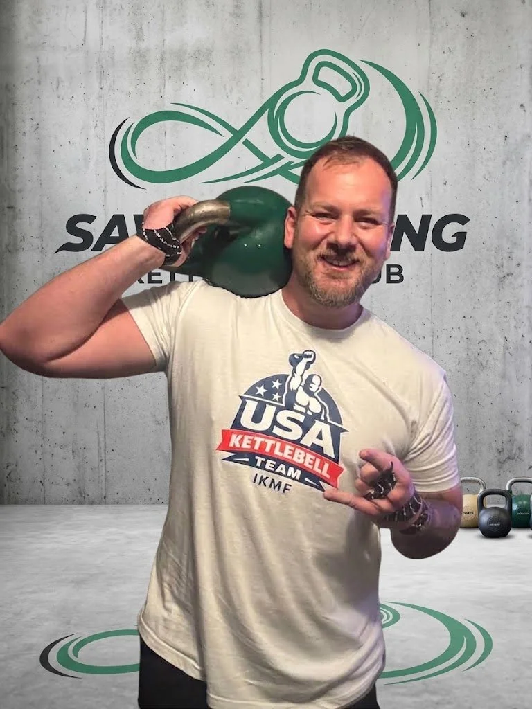 Smiling kettlebell sport athlete representing Team USA, holding a green kettlebell on his shoulder in front of a gym logo wall.