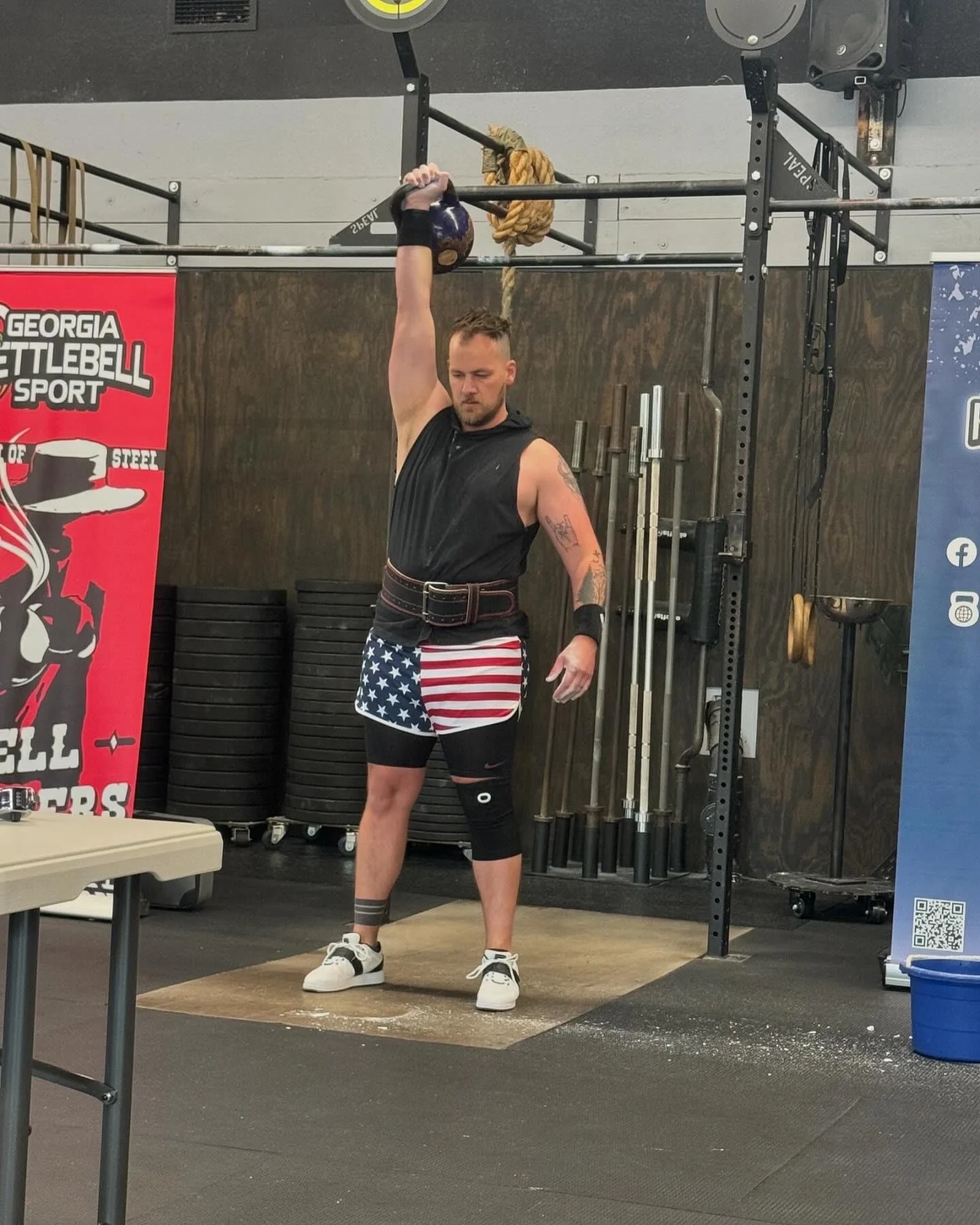 Kettlebell sport athlete wearing a lifting belt and American flag shorts performing a single-arm overhead kettlebell lift on a chalk-covered lifting platform.