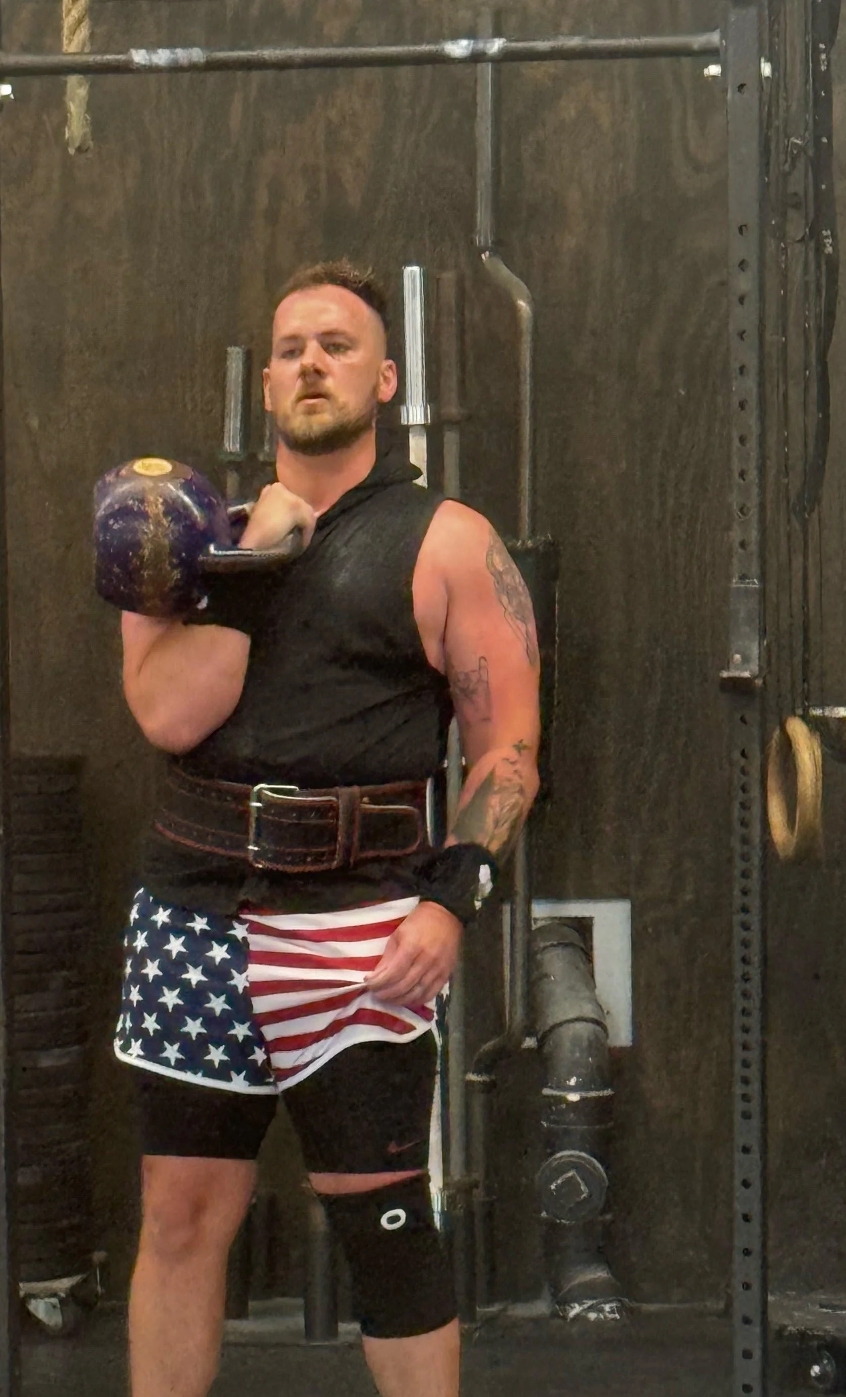 Male kettlebell sport athlete demonstrating the single-arm rack position with a chalked competition kettlebell, wearing a heavy lifting belt and American flag shorts.
