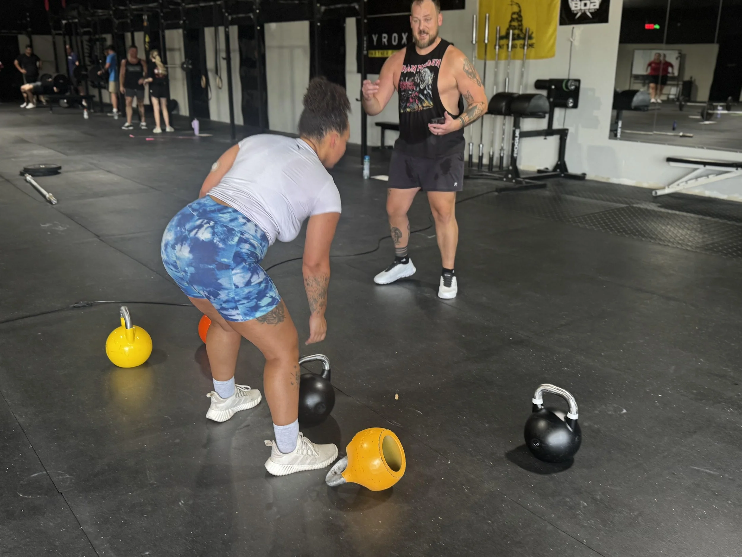 Kettlebell sport coach providing hands-on instruction to a female athlete setting up for a hardstyle lift on a rubber gym floor.