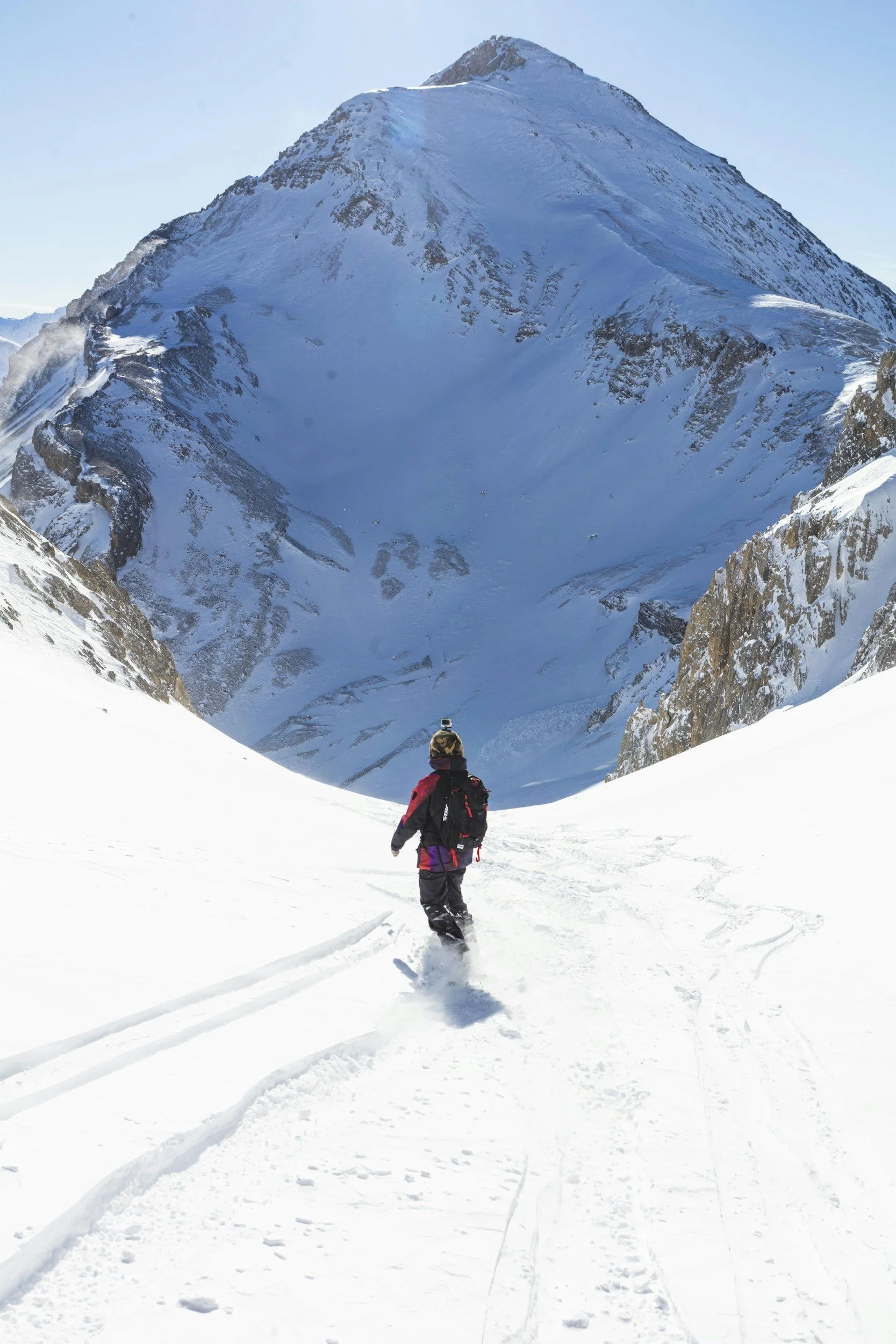 A person snowboarding through a snowy mountain pass with a large mountain in the background.