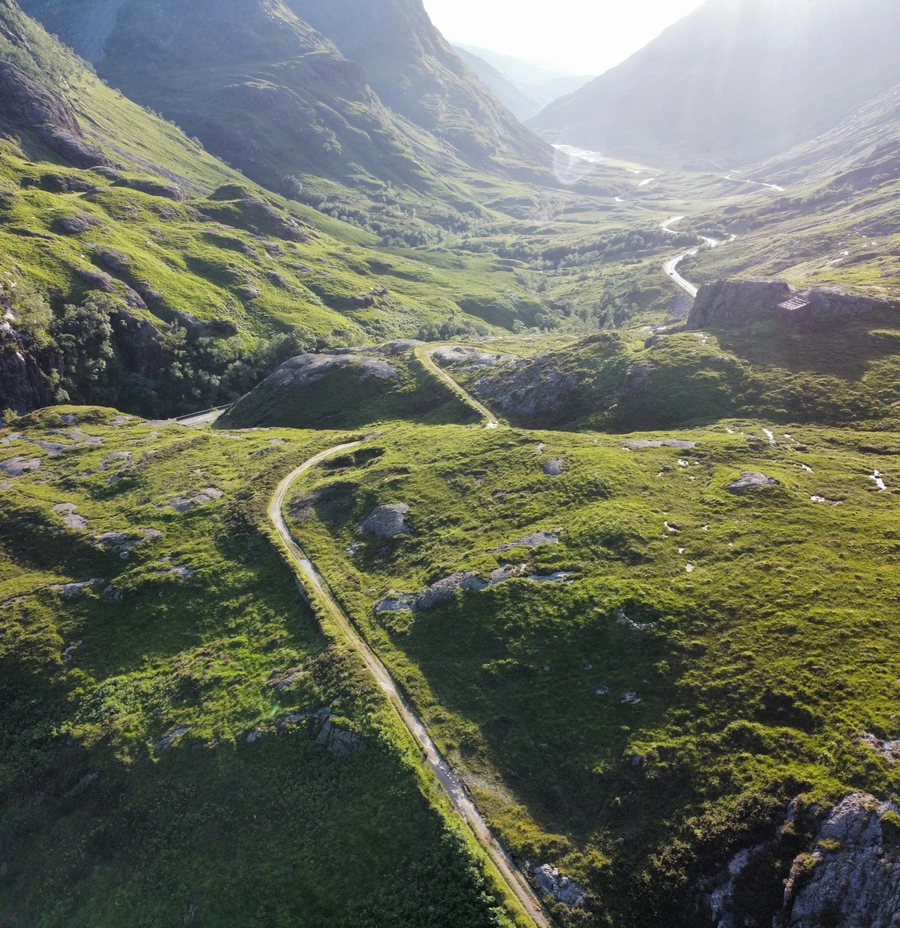 Aerial view of a winding road in a lush green mountain valley with steep hills and rocky outcrops, bright sunlight streaming over the landscape.