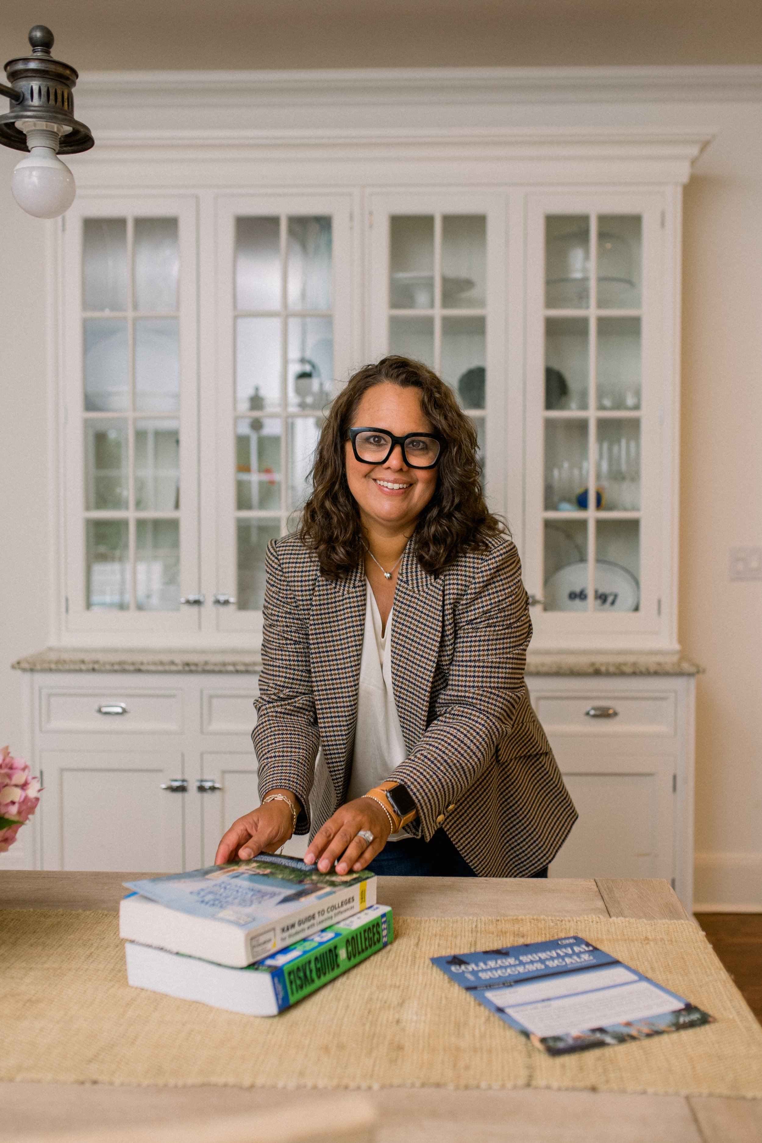 Woman smiling at a table with books about college guides, wearing glasses, a plaid blazer, and a smartwatch, in a room with white cabinet in the background.