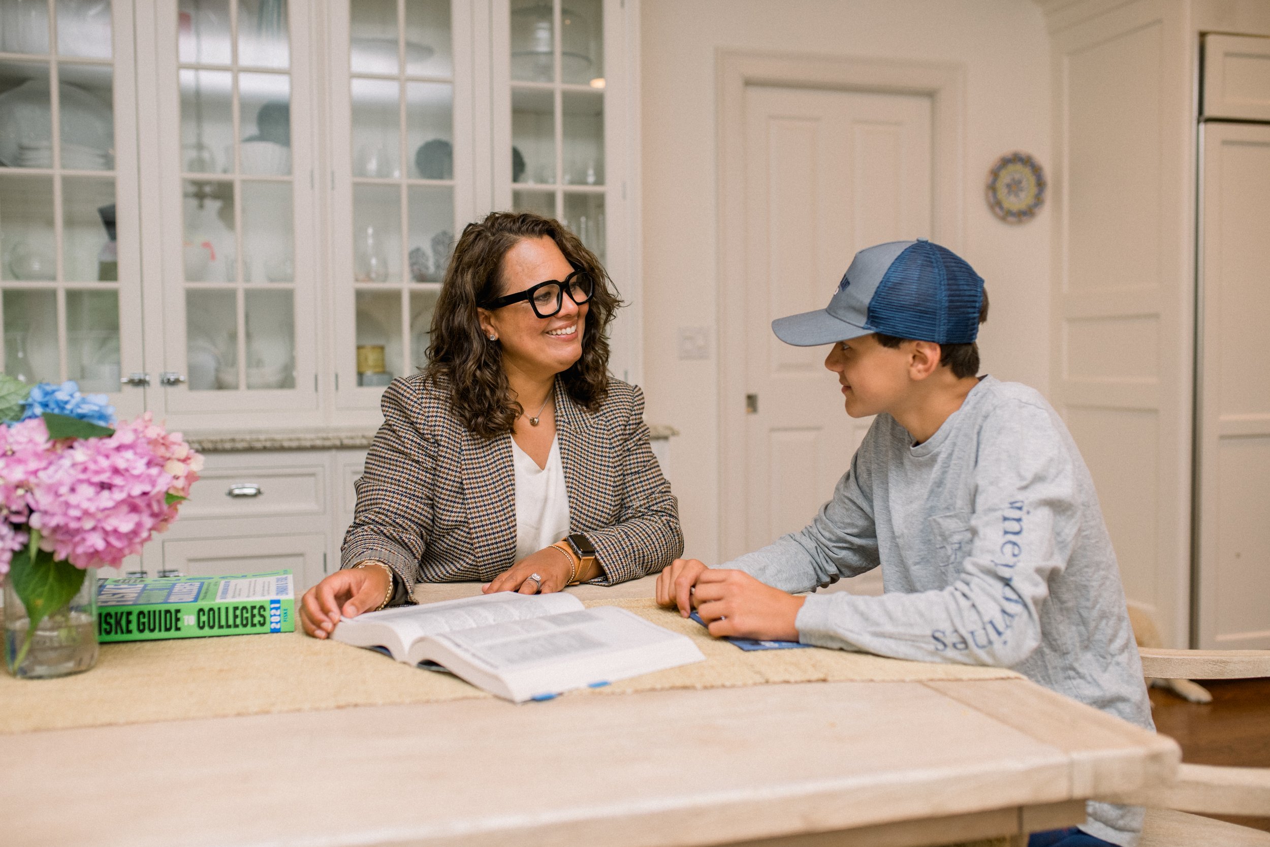 A woman with curly hair, glasses, and a blazer sitting at a kitchen counter, smiling, as she interacts with a boy wearing a baseball cap and sweatshirt. A book titled 'The Spectrum Guide to Colleges' and a vase of pink flowers are on the counter.