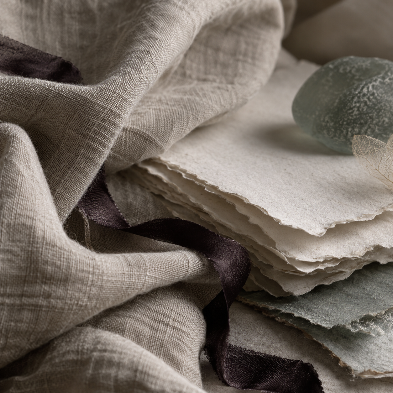 Still life of textured linen fabric, handmade paper, a smooth stone, and delicate leaves in neutral tones.