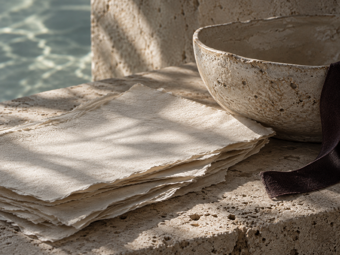 Stack of handmade paper next to a large speckled bowl, with a dark cloth, on a stone surface near water.