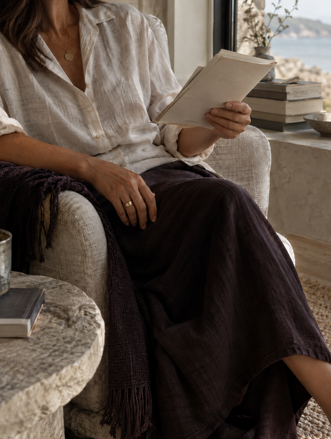 A woman sitting on a beige armchair near a window, reading a stack of papers. There are books and a ceramic vase with flowers on the windowsill.
