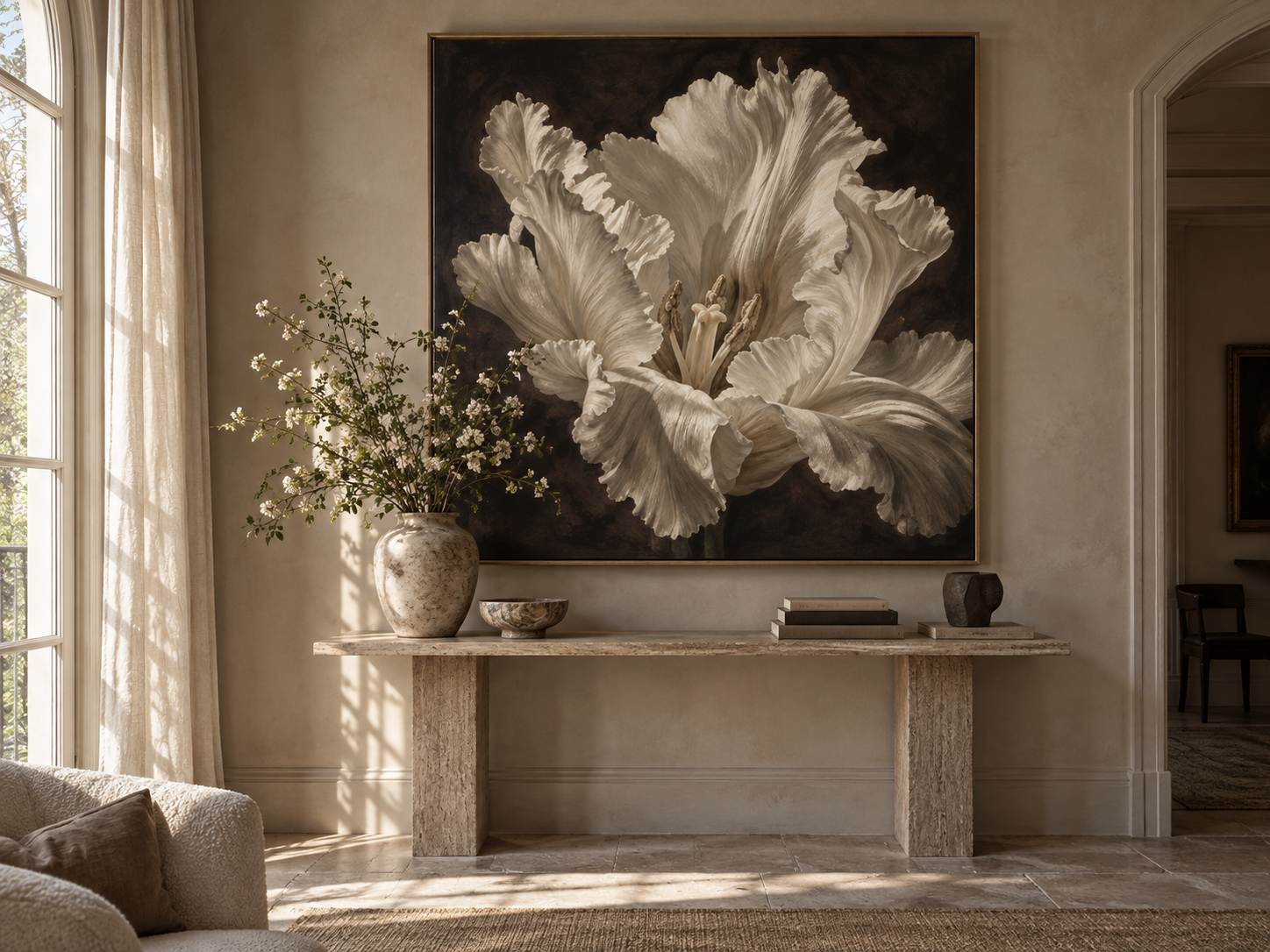 Elegant beige wall with large black, white, and gray floral painting above a marble console table. The table holds a tall vase with white flowers, a small bowl, a stack of books, and a sculpture. Light streams through a window with cream curtains to the left.
