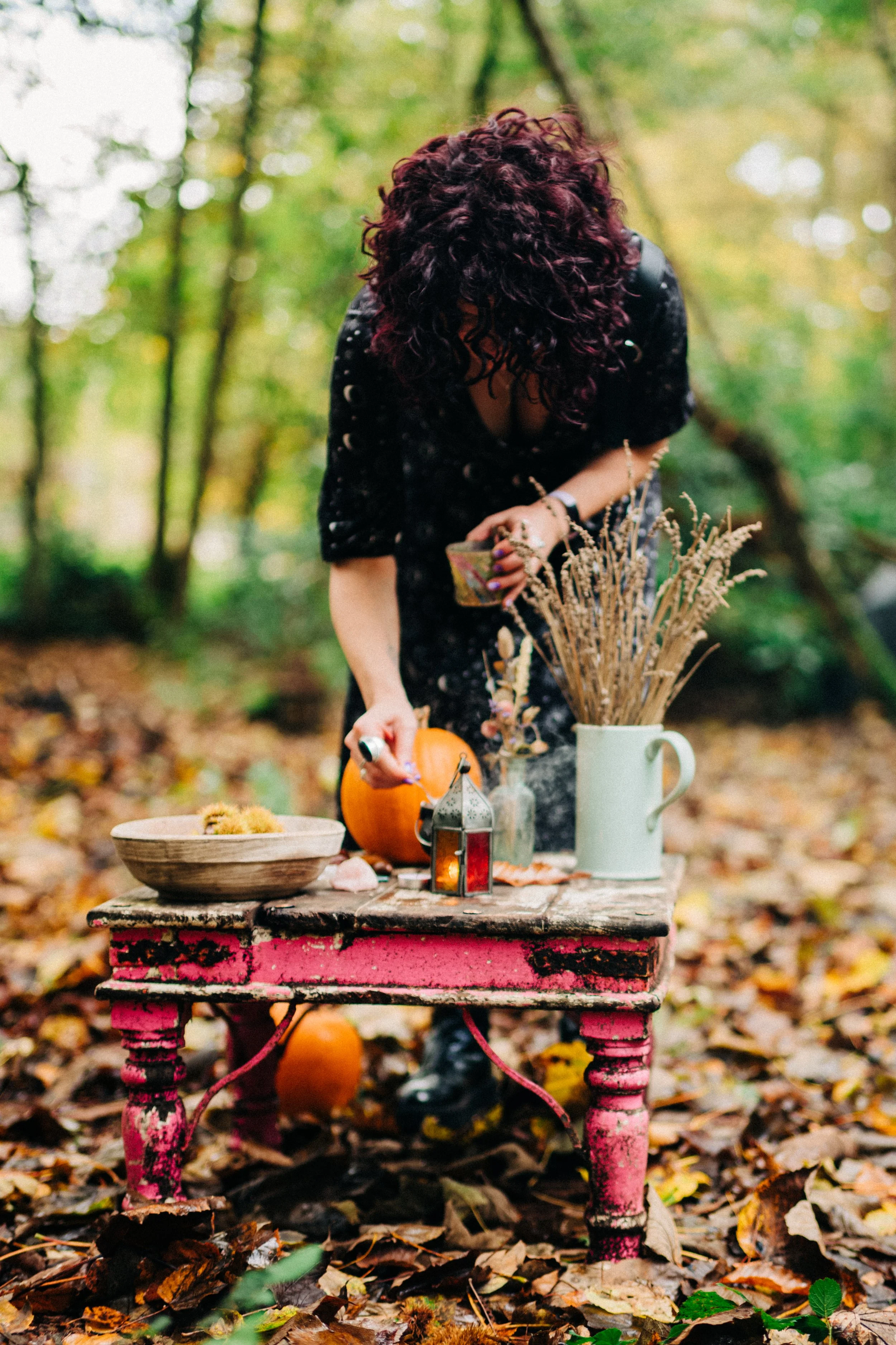 Person with curly hair and black witchy dress arranging autumnal decorations on a small pink painted wooden table outdoors, surrounded by fallen leaves in a woodland setting.