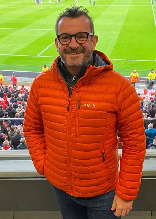 A man wearing glasses and an orange puffer jacket standing in a stadium, with a soccer field and spectators in the background.
