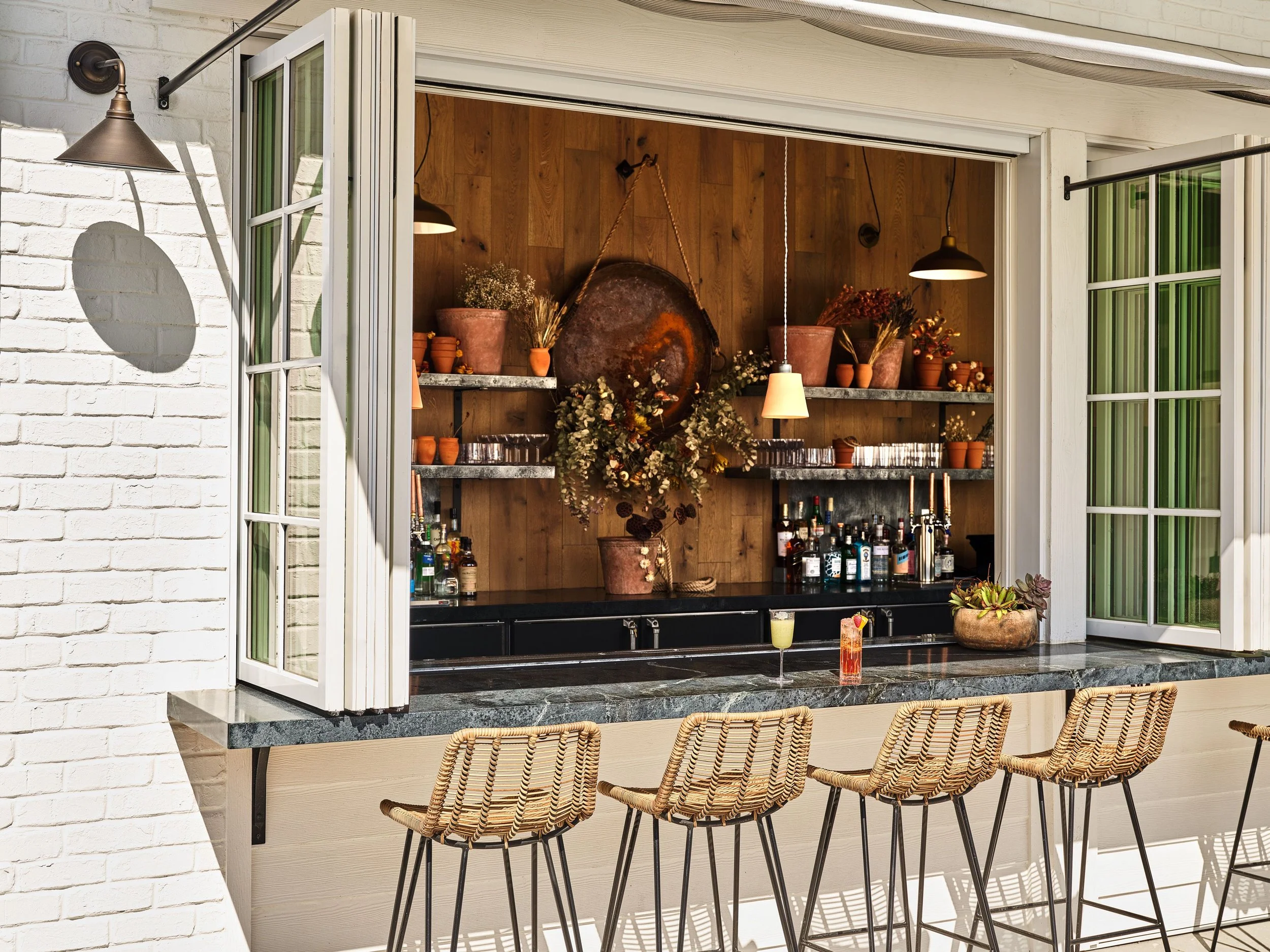 View of a stylish outdoor bar area with a marble countertop, surrounded by rattan bar stools, set against a white brick wall with open glass doors revealing a cozy indoor space with wooden paneling, potted plants, bar bottles, and warm lighting.