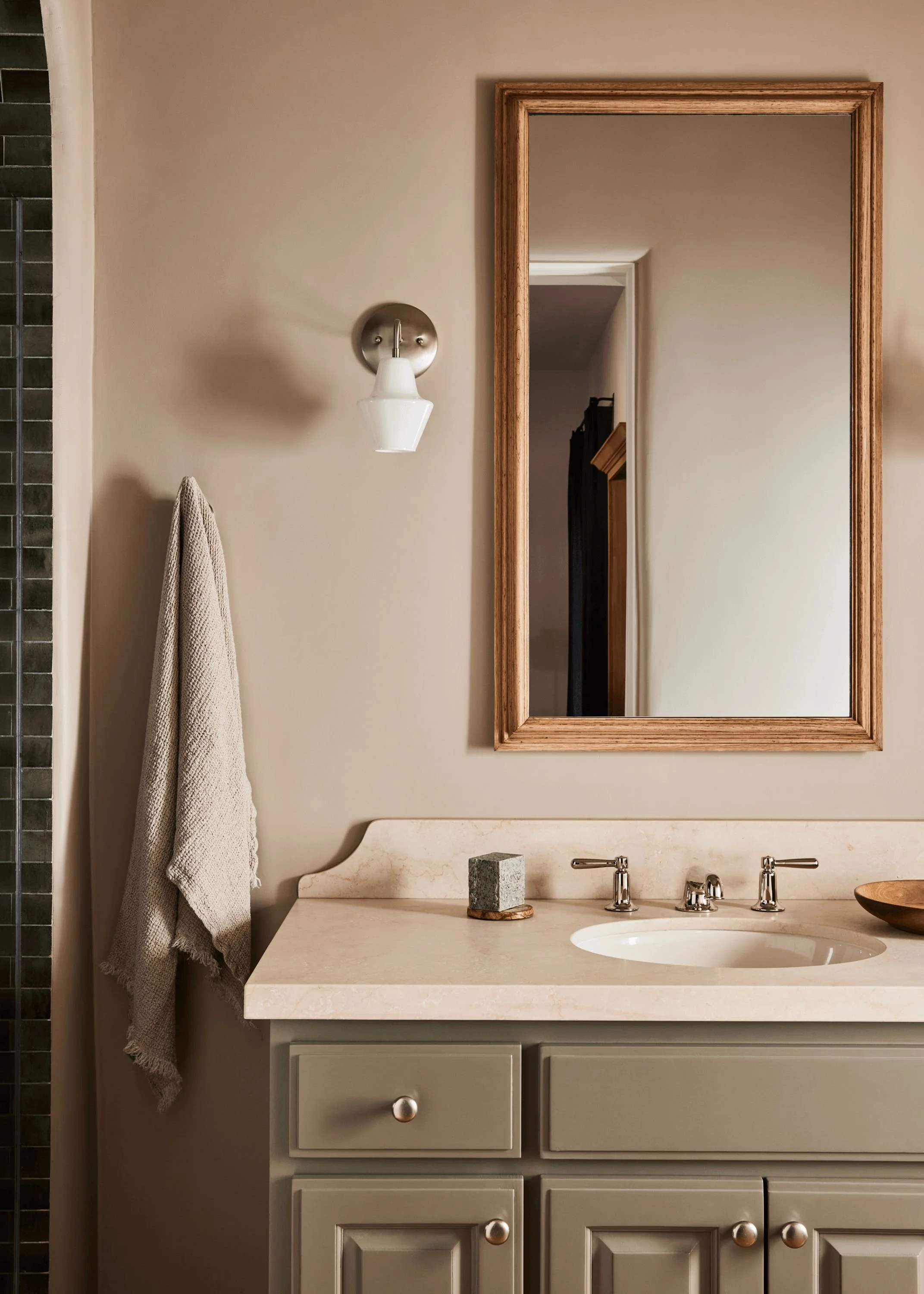 Bathroom vanity with a gray cabinet, marble countertop, oval sink, bronze faucet handles, a square stone accessory, and a large wooden framed mirror. To the left, a beige towel hangs on a hook. A wall-mounted white sconce light is to the left of the mirror.