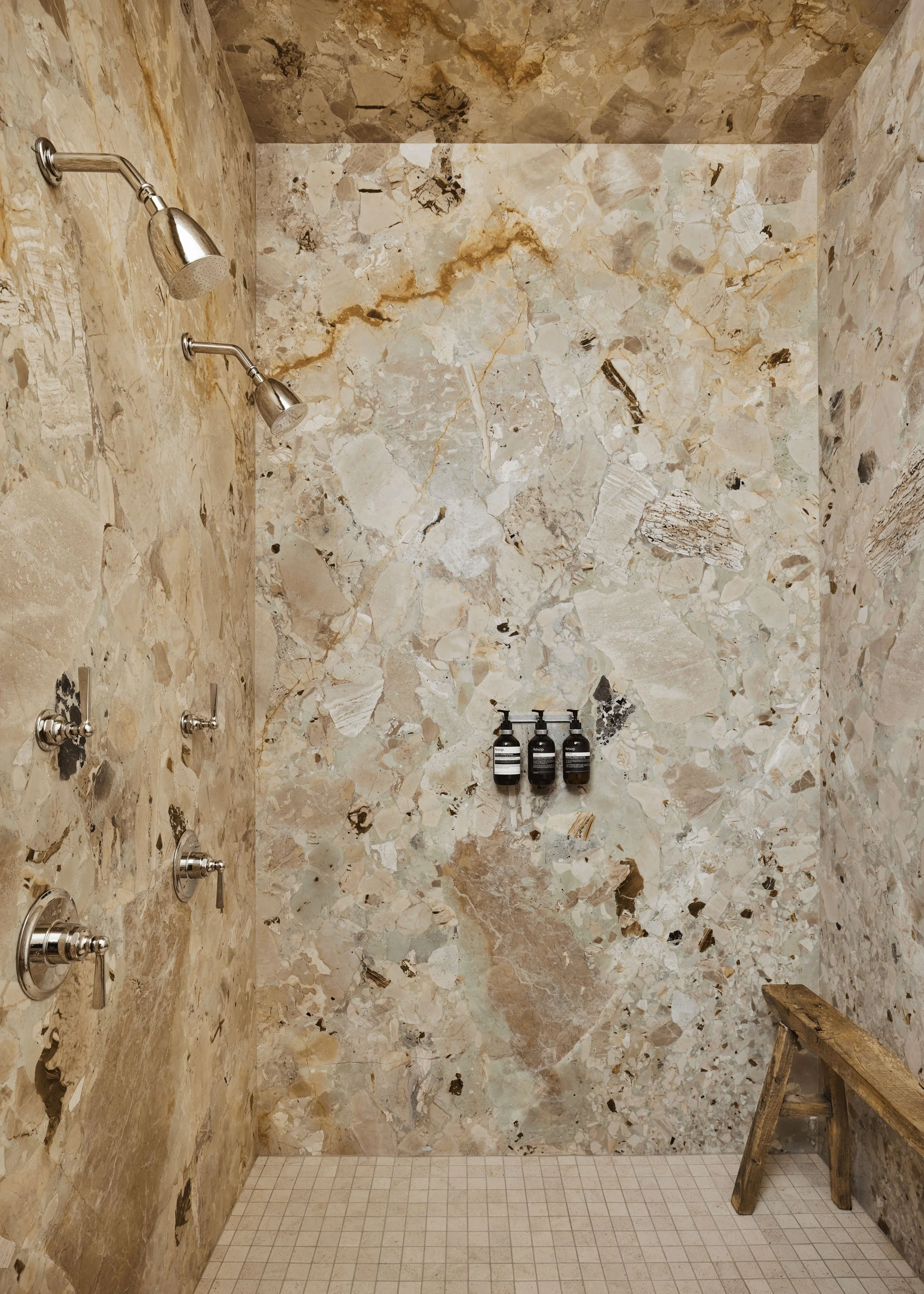 Stone tiled shower with two rainfall showerheads, three bottles of shower products on a ledge, an old wooden bench, and soap on the floor.
