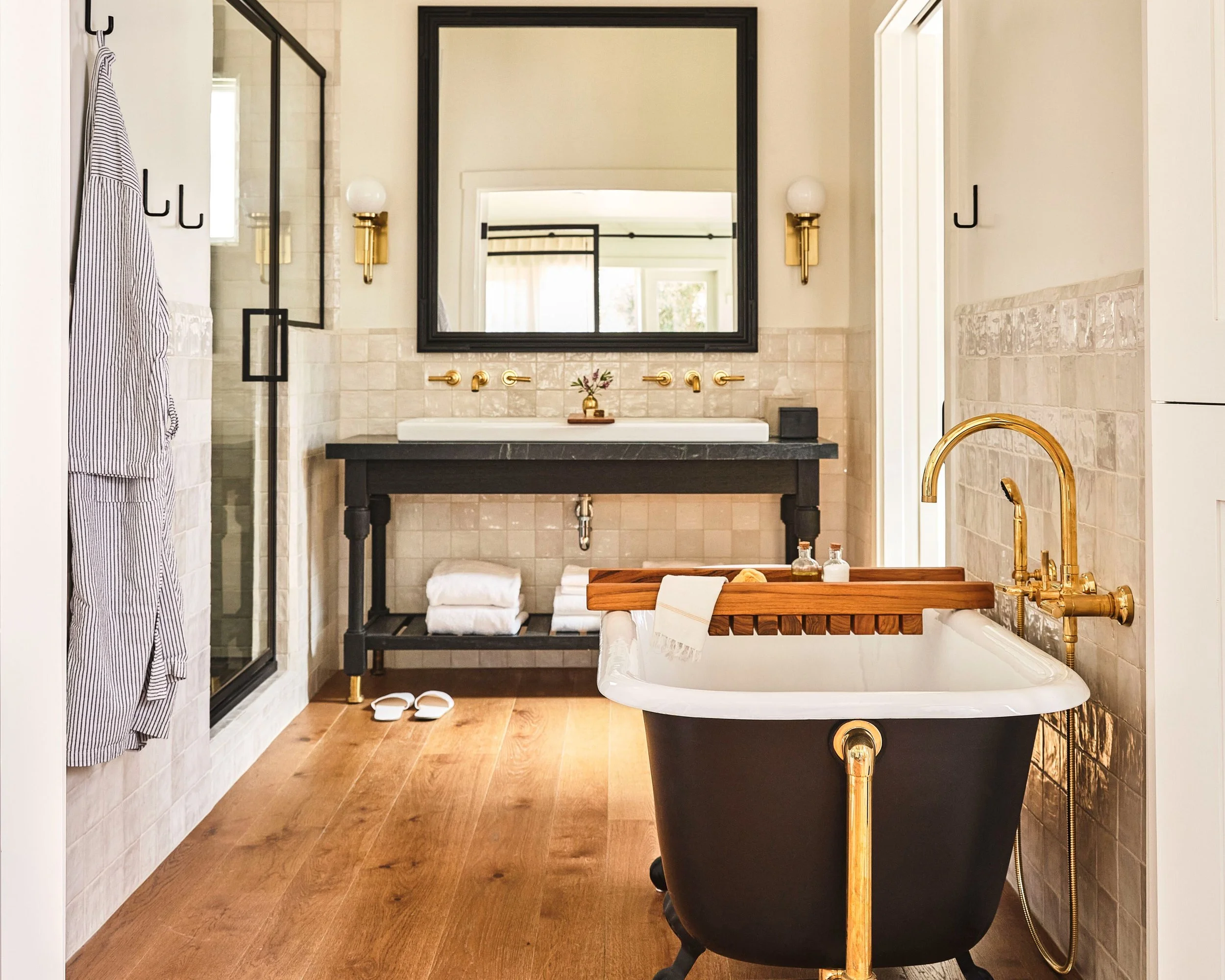 A modern bathroom with a large mirror, black and gold fixtures, a black bathtub with gold fixtures, a wooden tray with towels, white slippers, a towel hanging on a black hook, and a shower enclosure with a glass door.