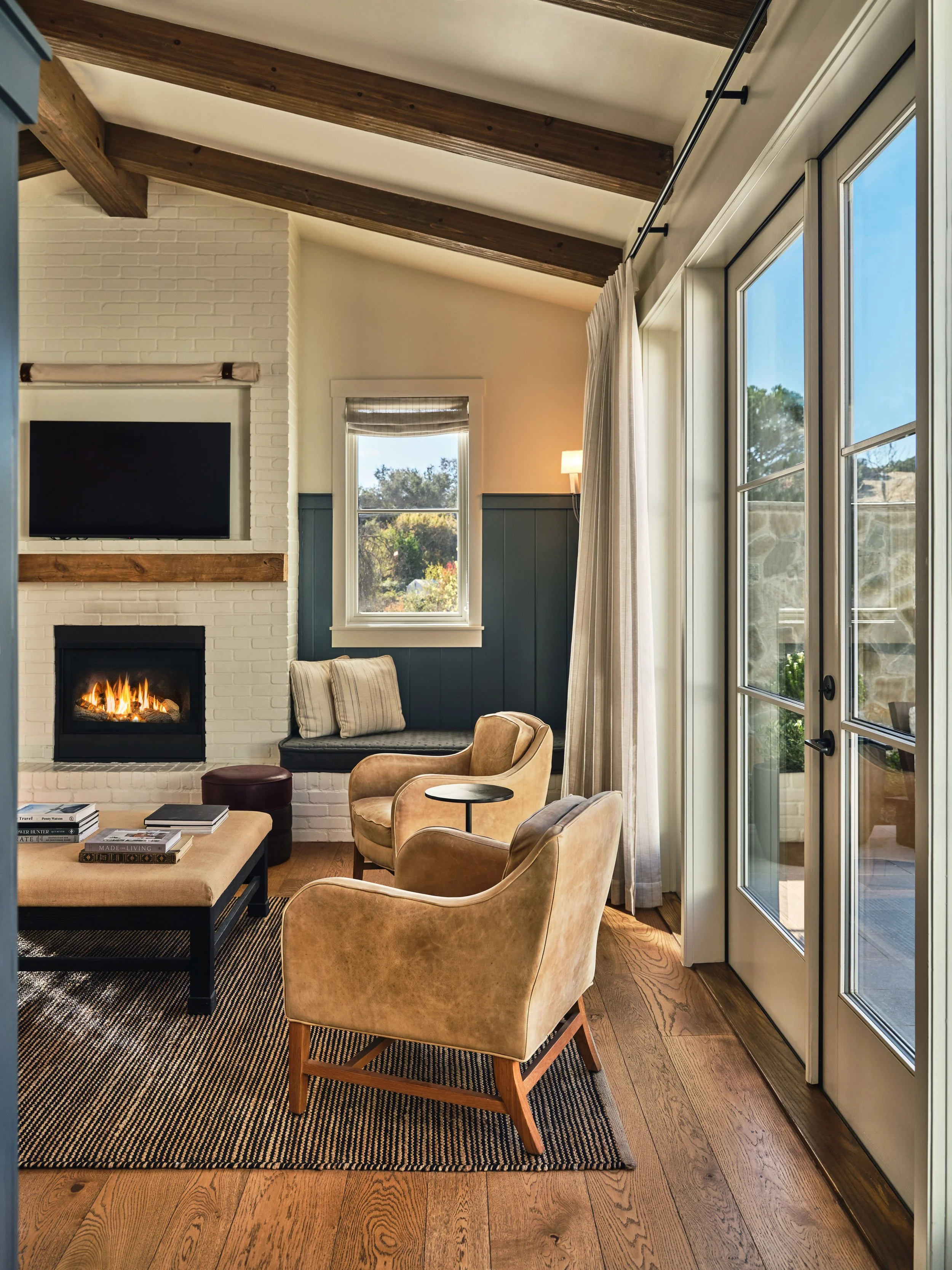 Living room with beige armchairs, a black coffee table, a pattern rug, a white brick fireplace with a fire, a wall-mounted TV, large windows, and wooden flooring and ceiling beams.