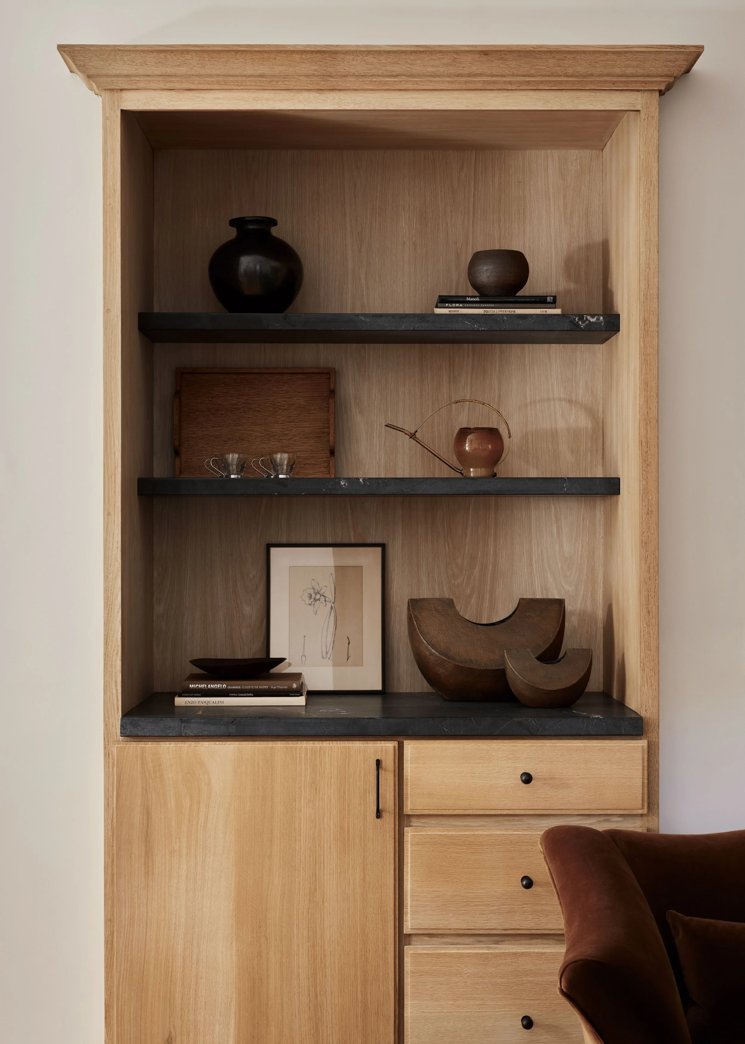 A wooden bookshelf with black marble shelves displaying vases, books, and decorative items, alongside a framed botanical print and a brown velvet armchair