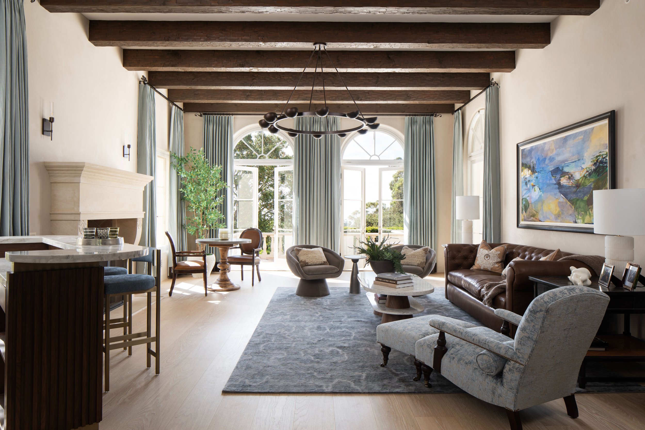 Living room with high ceiling, exposed wooden beams, large arched windows, and light-colored walls. The room features a brown leather sofa, patterned armchair, coffee table, abstract wall art, and various decorative items.