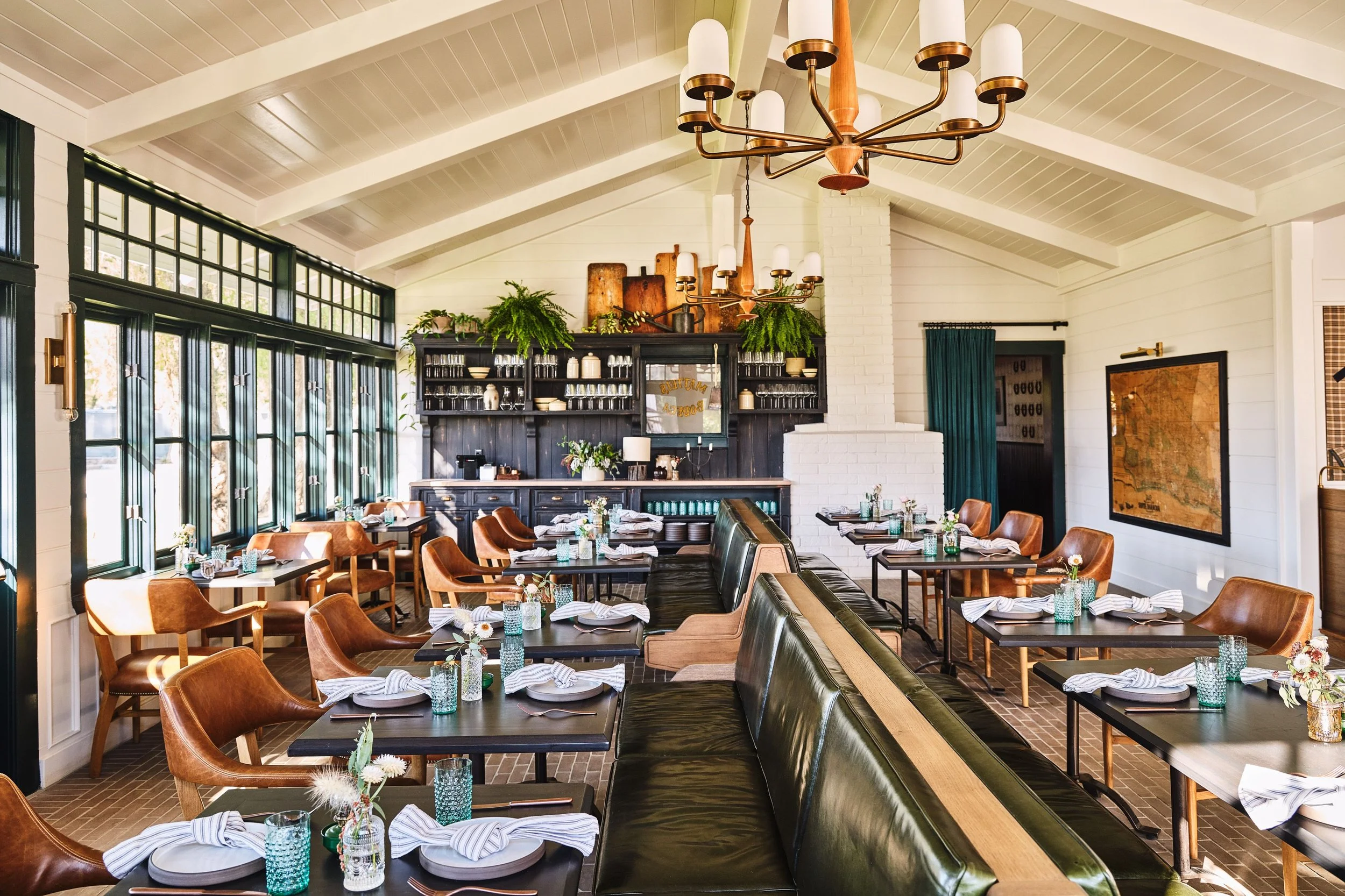 Interior of a restaurant with wooden tables and tan leather chairs, set with plates, napkins, and small vases of flowers. Large windows along one wall allow natural light, and a white brick fireplace behind a dark wooden shelf displays glassware and decorative objects. A brass chandelier hangs from the vaulted ceiling.