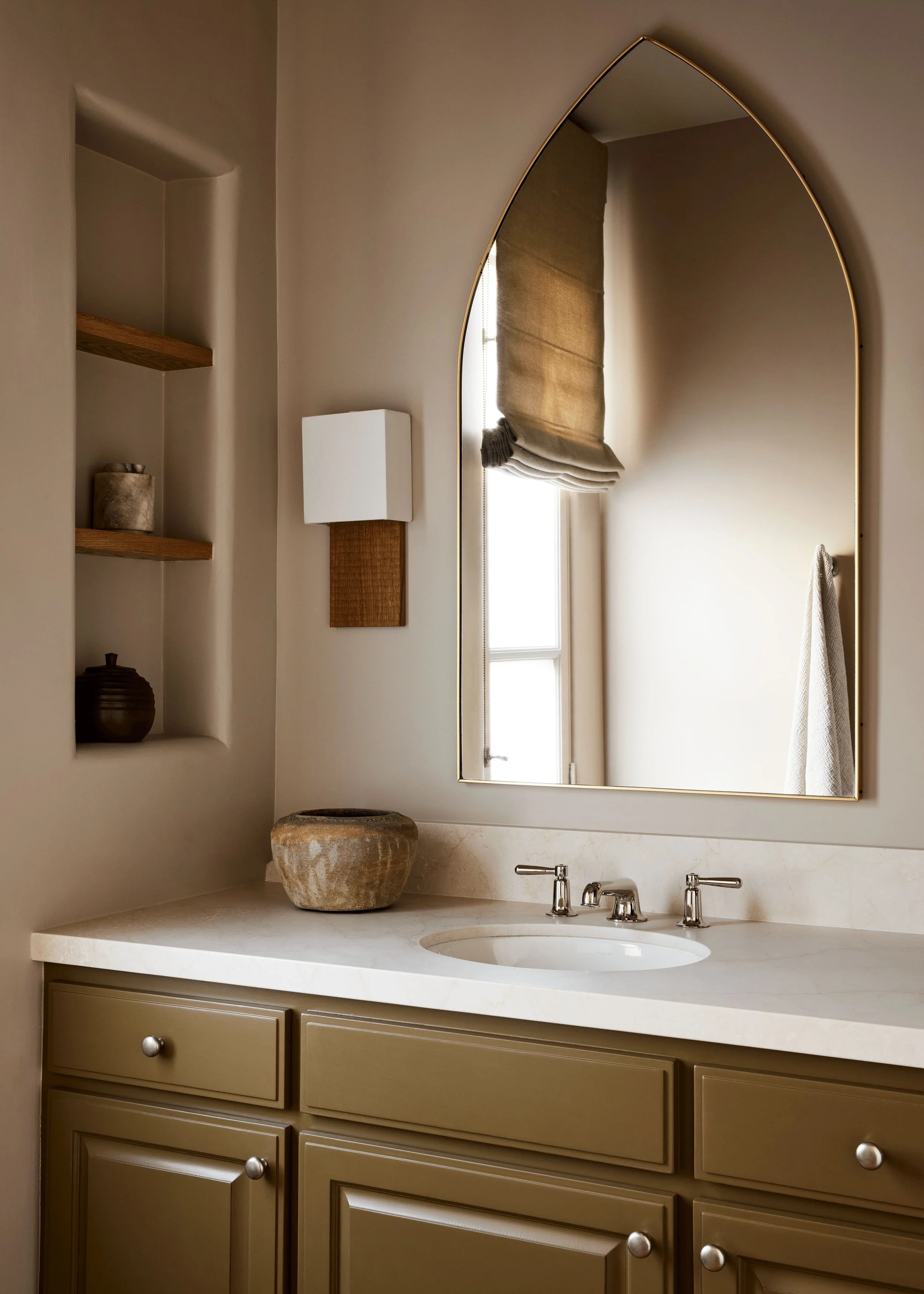 A bathroom vanity with a beige cabinet, a white countertop, and a white sink with silver fixtures. Above the sink is a large, arched mirror with a gold frame. To the left, there are open shelves with decorative items, and a window with a roll-up beige shade. The wall is painted light neutral color, and there is a small wall lamp beside the window. The countertop has a textured stone vessel and a small woven basket.