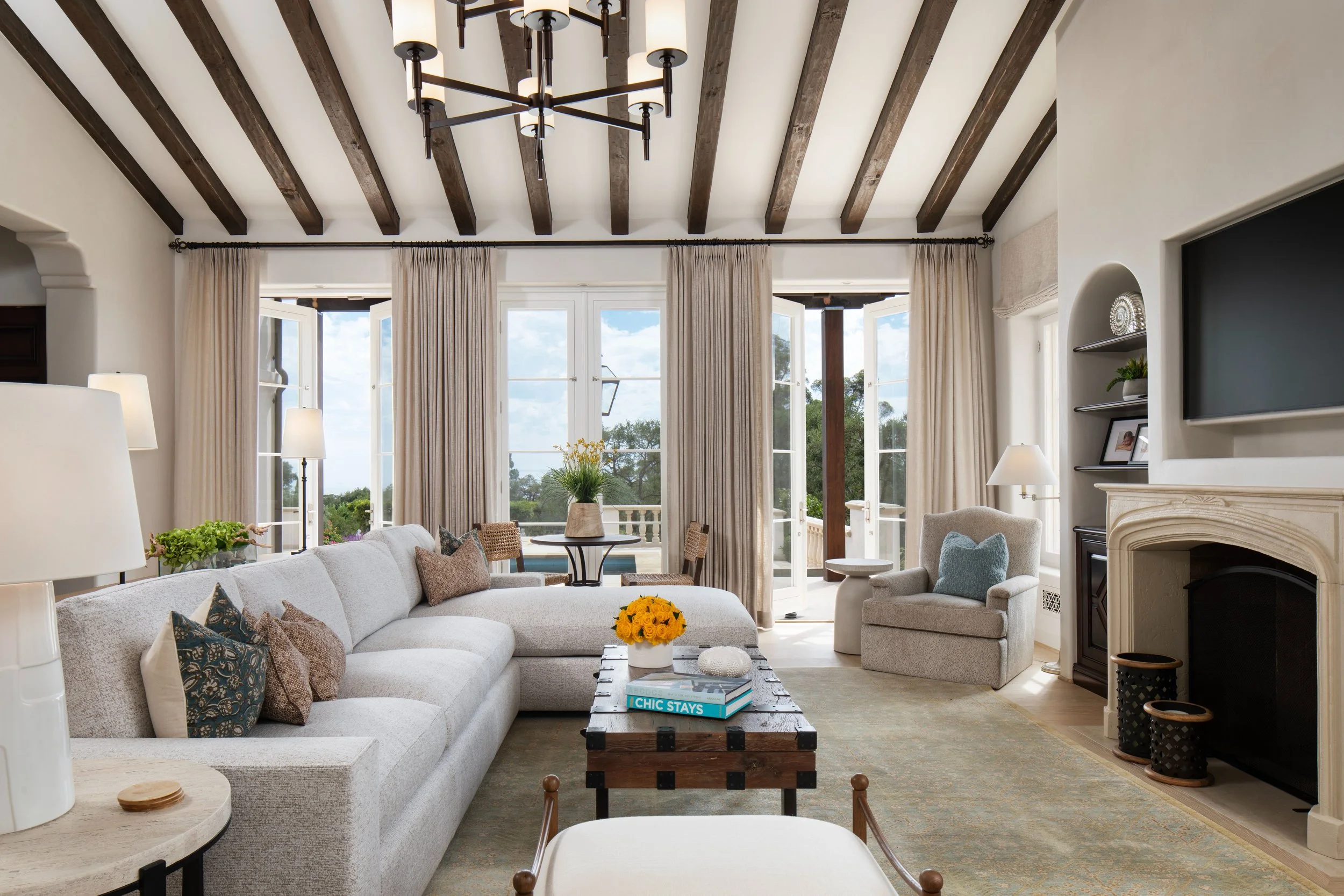 Living room with large windows, white sofa, armchair, coffee table with books, decorative pillows, fireplace, and curtains.