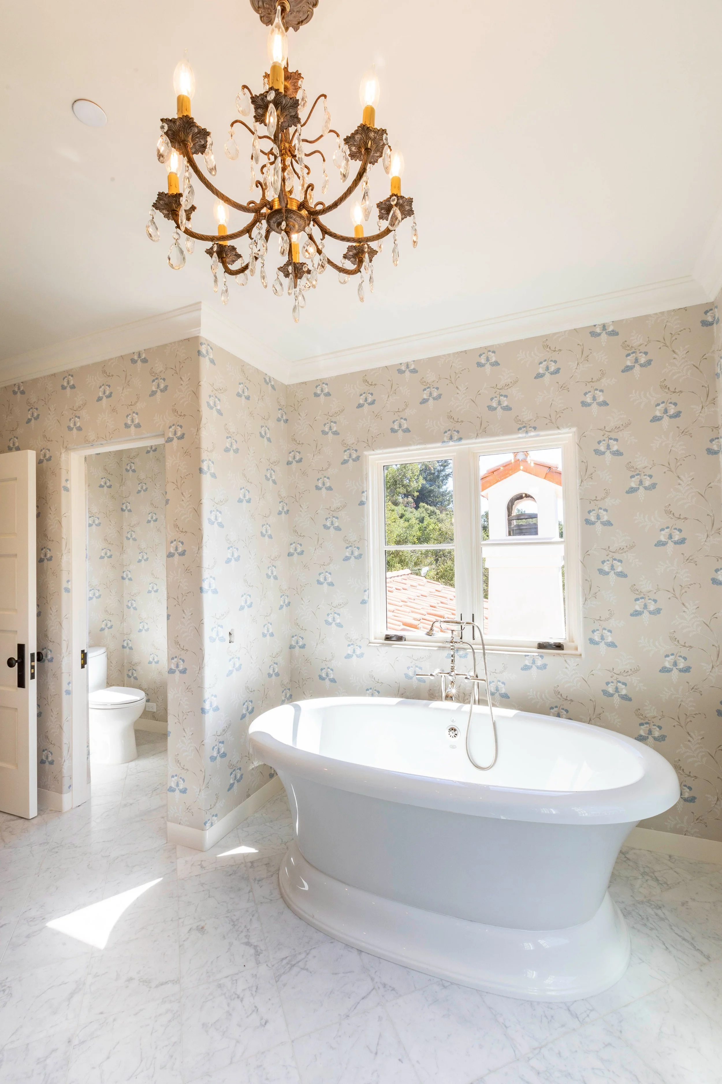 An elegant bathroom with a freestanding bathtub beneath a window, floral wallpaper, and a chandelier hanging from the ceiling.