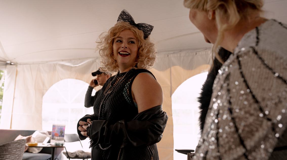 Two women in shiny, stylish outfits inside a light-colored event tent, with one woman smiling at the camera and the other looking away.