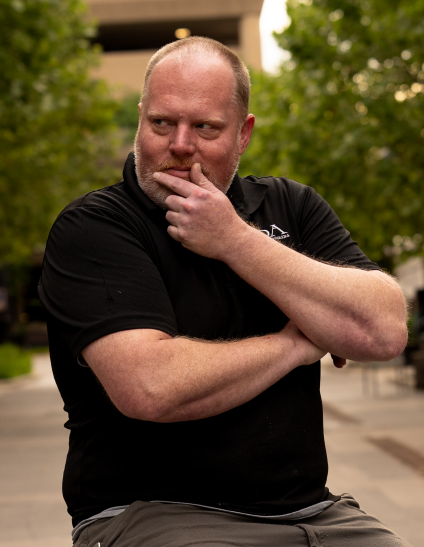 A man with a beard and short hair, wearing a black shirt, standing outdoors with greenery in the background, looking thoughtful with his hand on his chin.