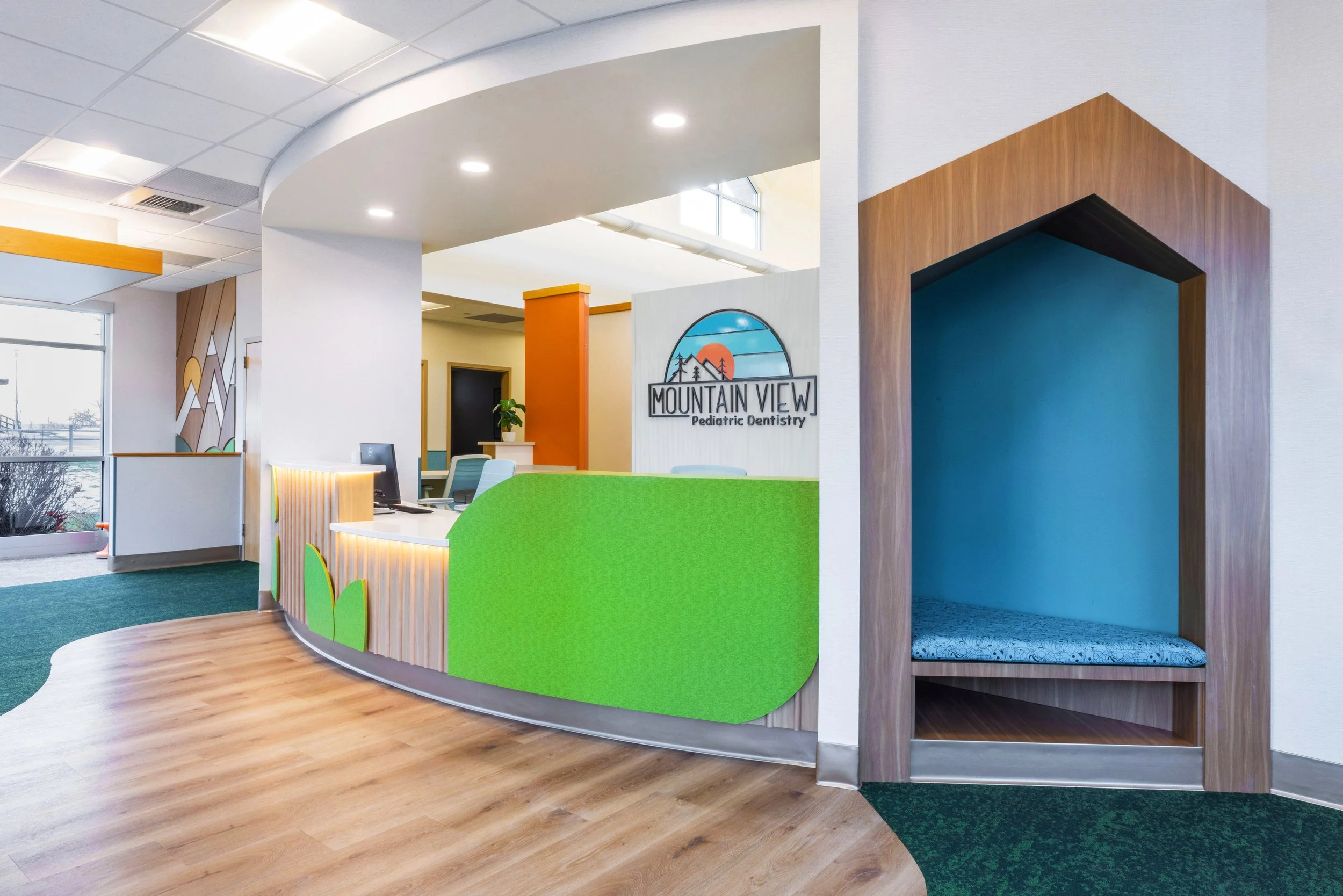 Reception area of Mountain View Pediatric Dentistry with a curved green counter, a small seating nook with a blue cushion, and a sign featuring a landscape with mountains, trees, and a sunset.
