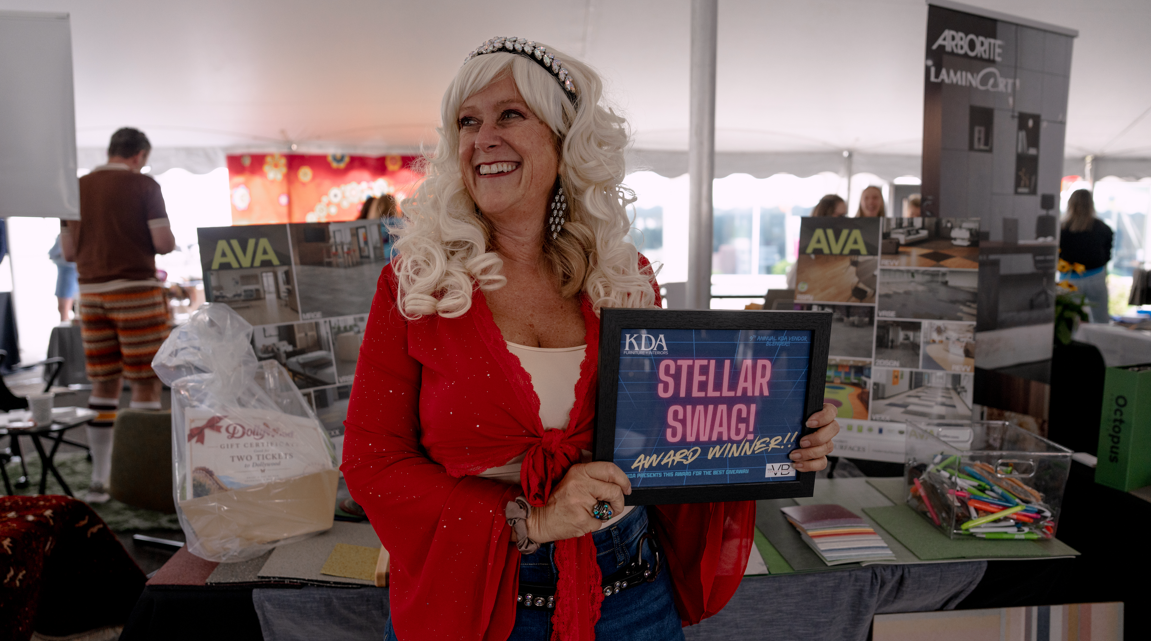 A woman with long curly blonde hair, wearing a red blazer and a headband, smiling and holding an award plaque that reads "STELLAR SWAG! AWARD WINNER!" at an indoor event with booths and other people in the background.