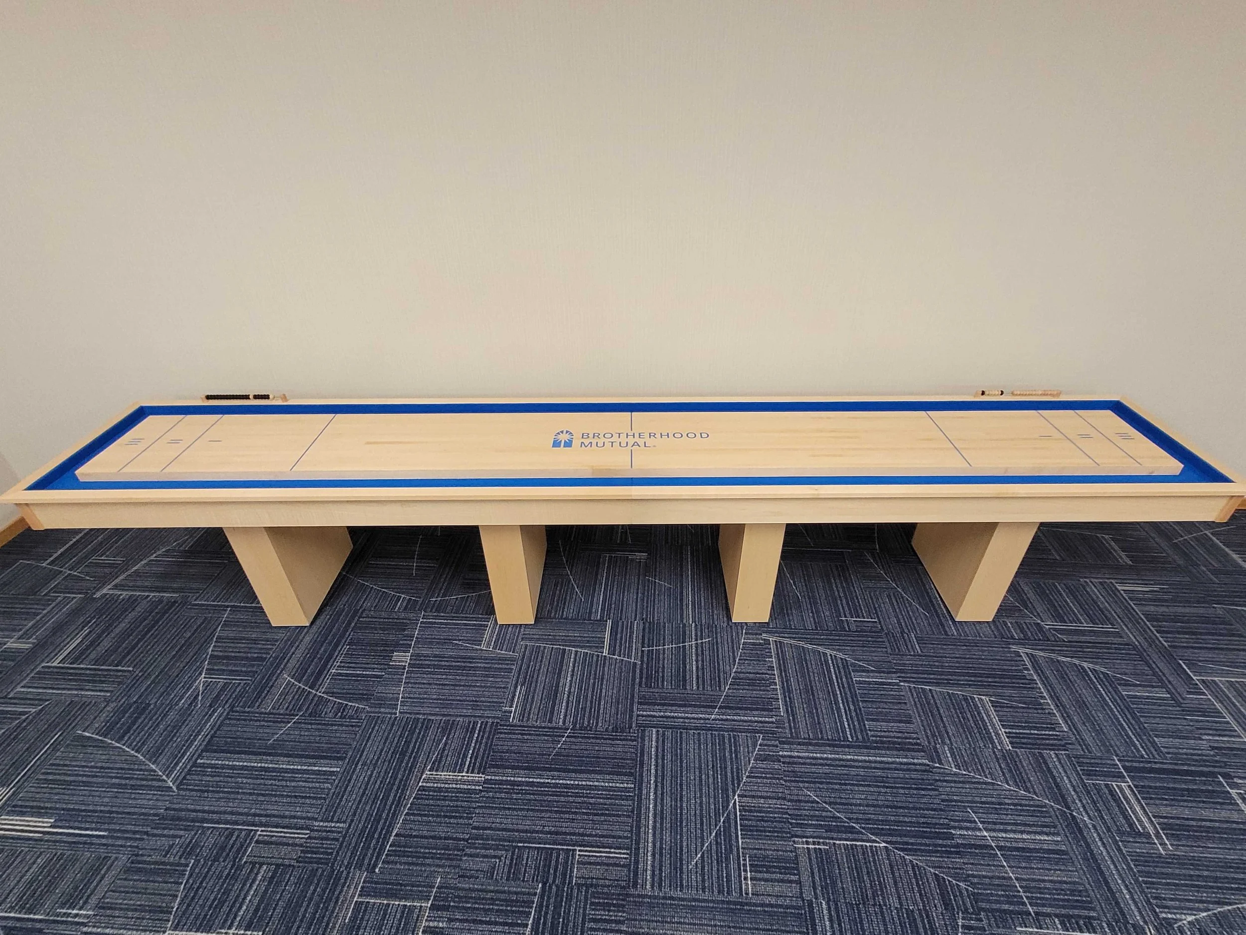 A shuffleboard table with a blue border, featuring the logo of Brotherhood Mutual, is positioned against a plain wall on a dark geometric patterned floor.