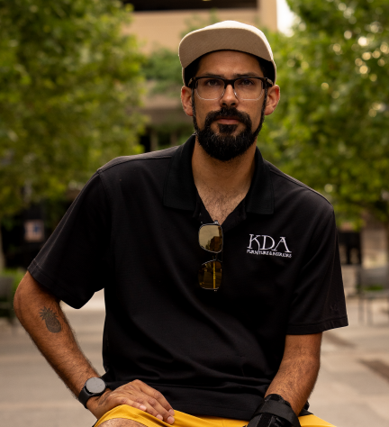 Man with a beard and glasses wearing a black polo shirt with 'KDA' logo, beige cap, and sunglasses hanging from shirt, sitting outdoors with trees and a building in the background.