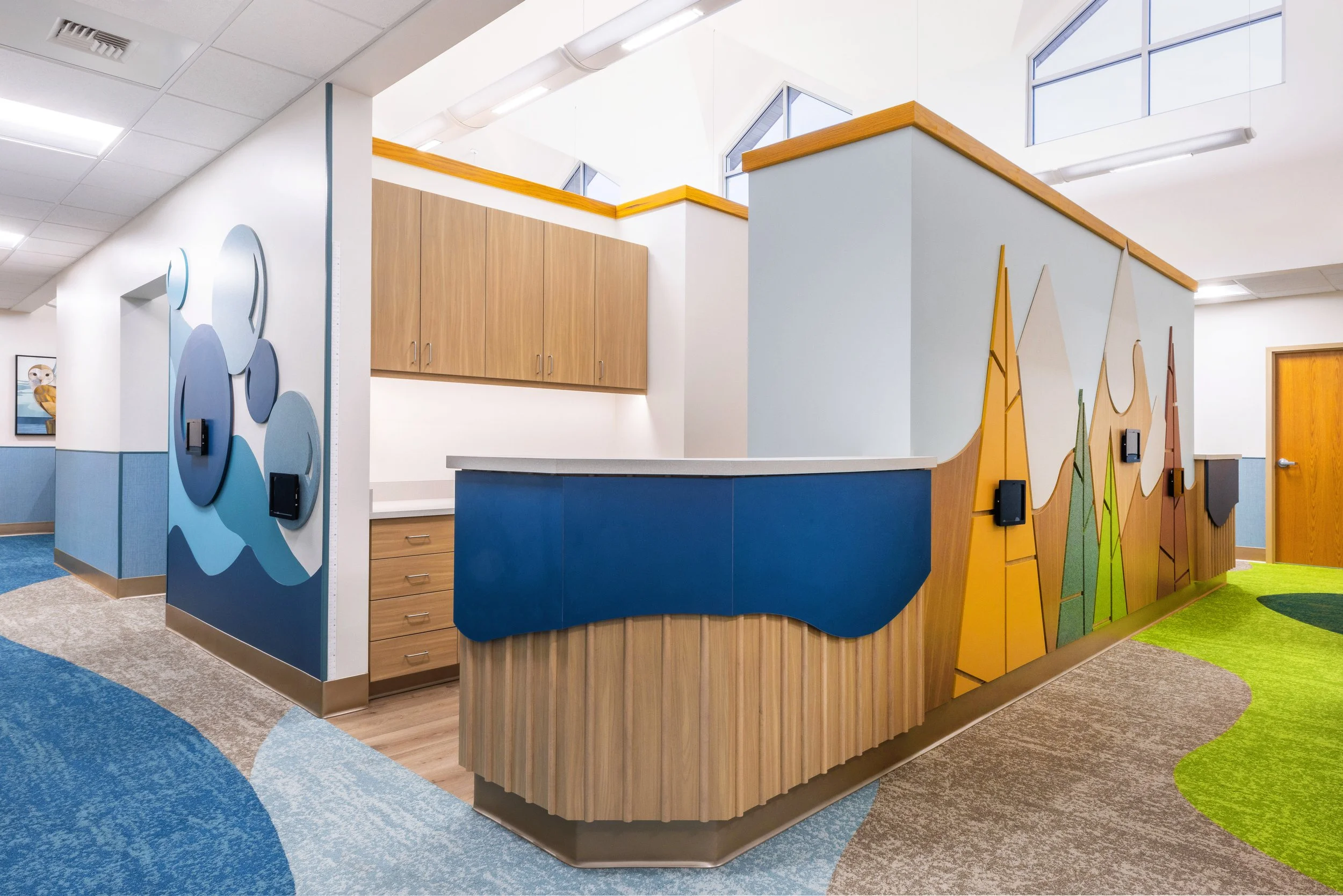 Colorful indoor space with artistic wall designs, wooden cabinets, and a curved reception desk, suggesting a child-friendly environment such as a preschool or pediatric clinic.