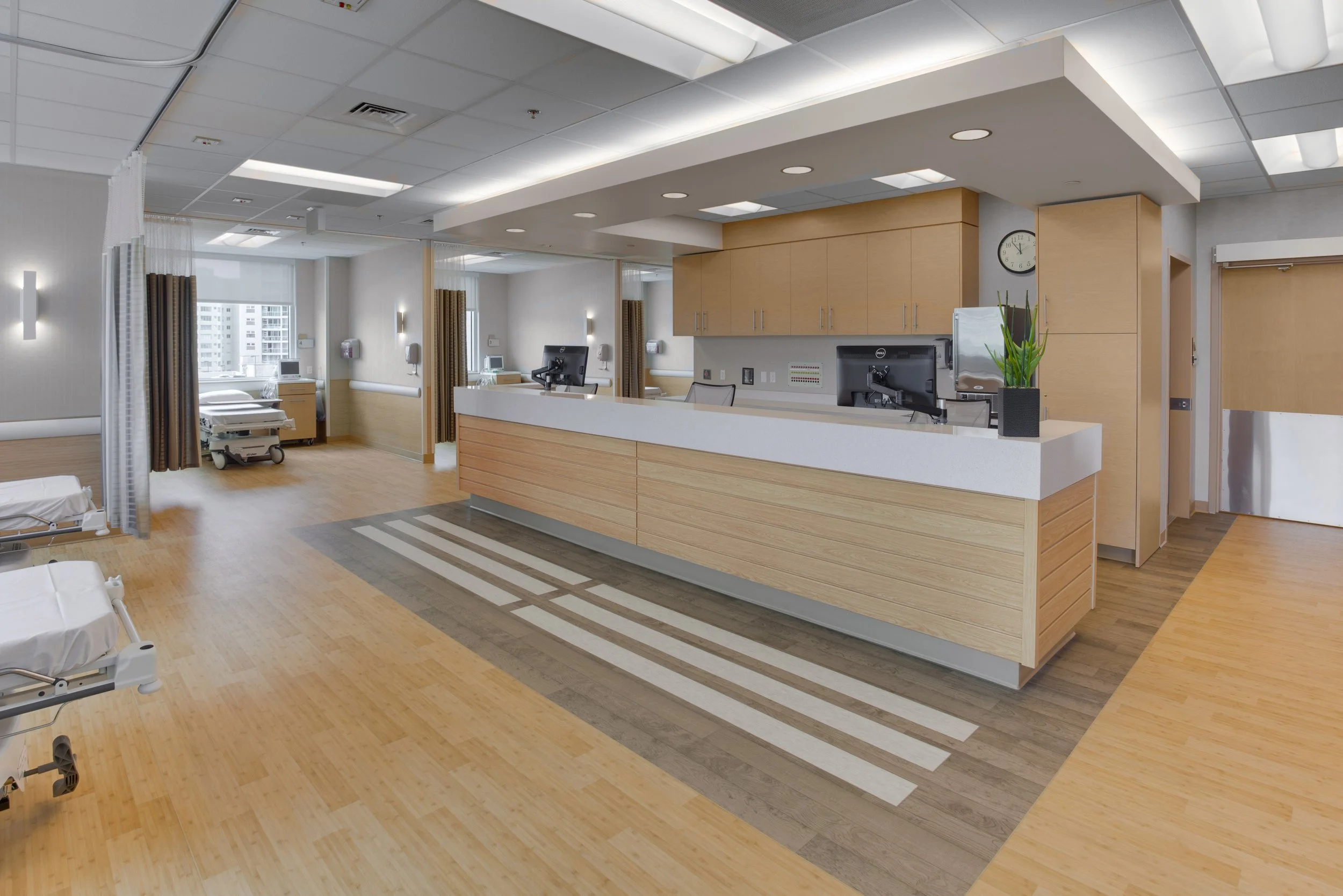 A hospital reception area with a wooden counter, computers, a potted plant, a wall clock, and hospital beds visible in the background. Bright lighting and large windows with city buildings outside.