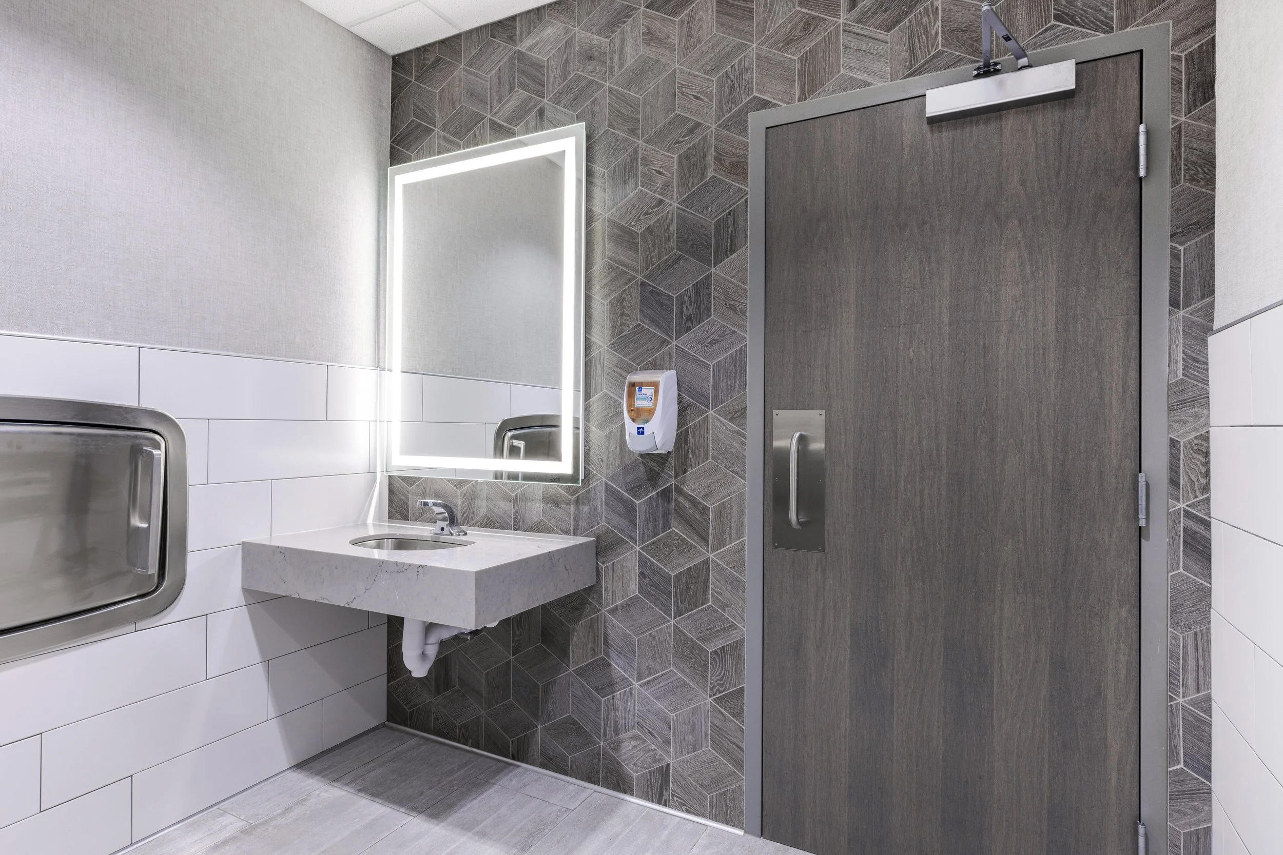 Restroom with a mirror lit with LED lighting, a small sink, a soap dispenser, and a closed wooden door with a metal handle and a latch.