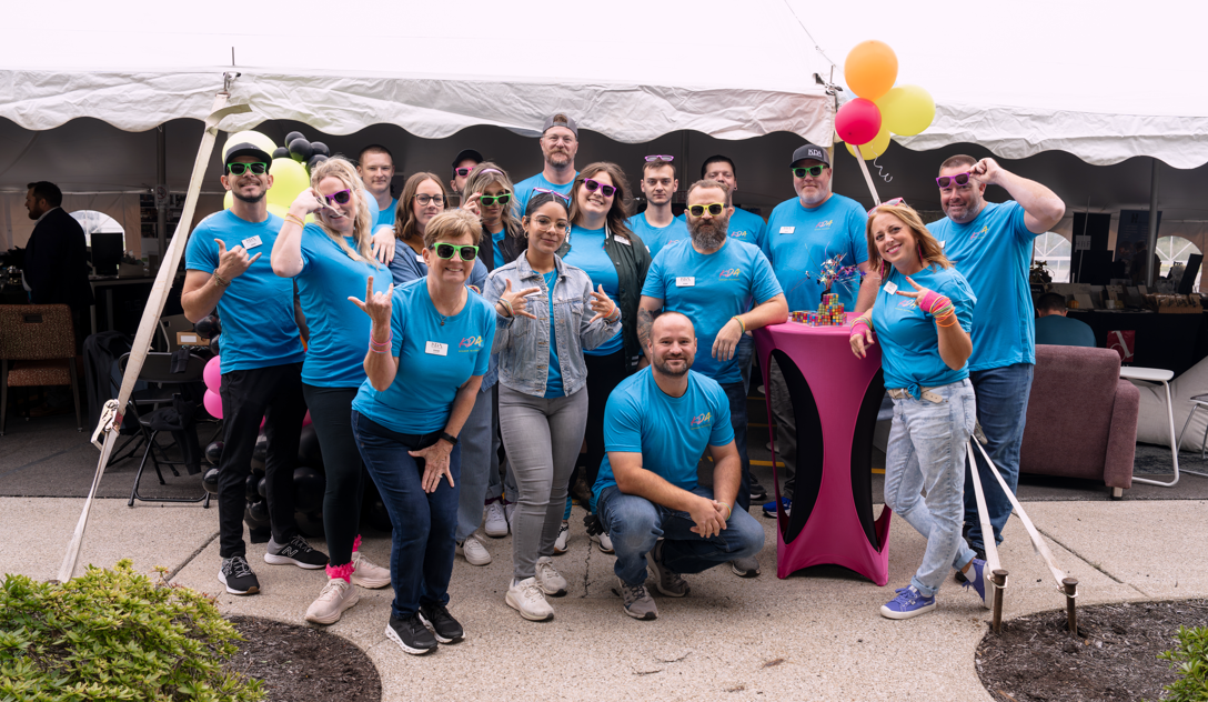 Group of people wearing blue T-shirts posing for a photo under a white tent with colorful balloons and a pink table with decorations.