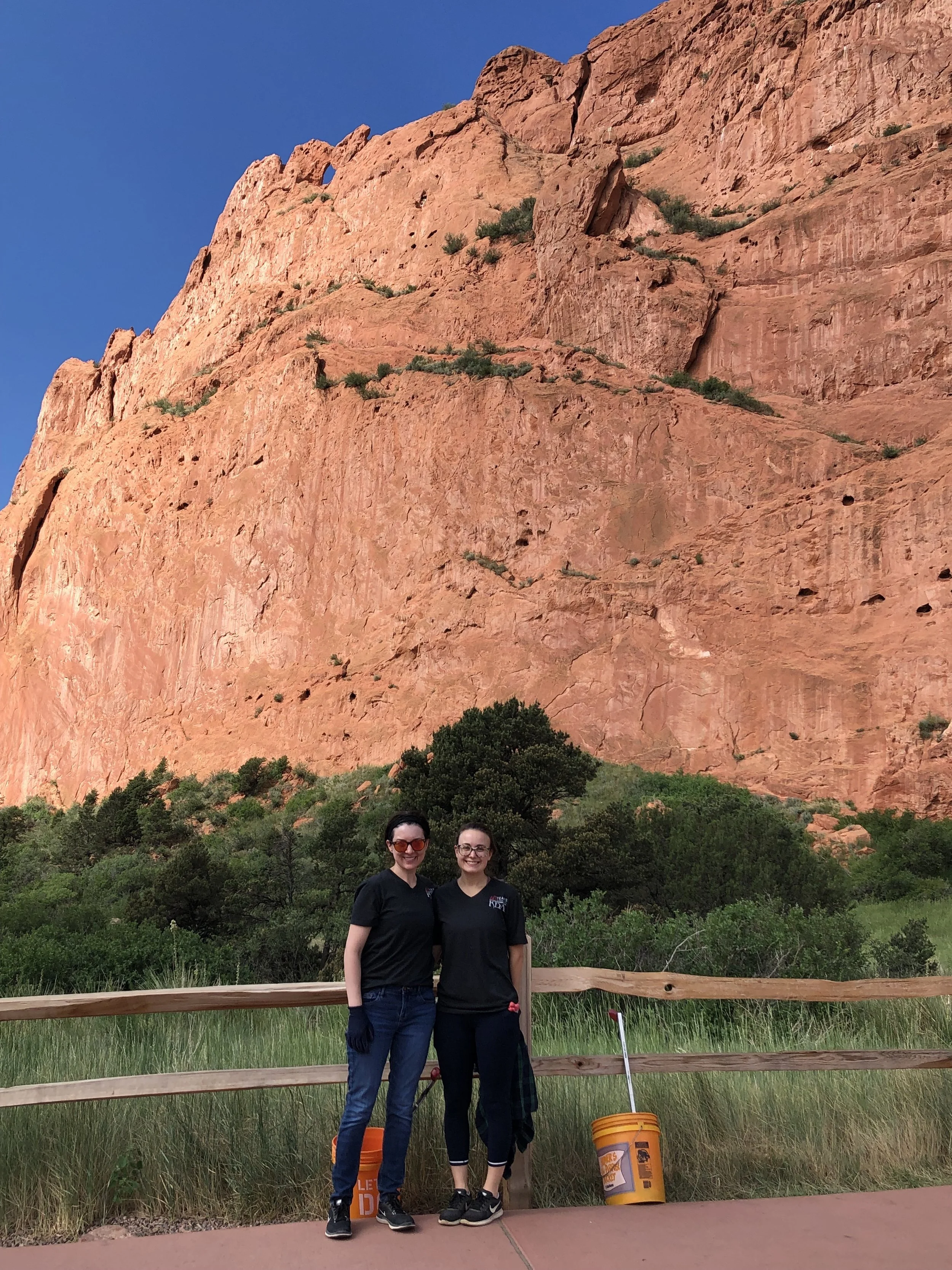 Two women standing outdoors with a large red rock formation in the background, trees, a wooden fence, and orange buckets by their feet.