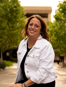 A woman with brown hair smiling, wearing a white jacket and black shirt, outdoors with trees and a building in the background.