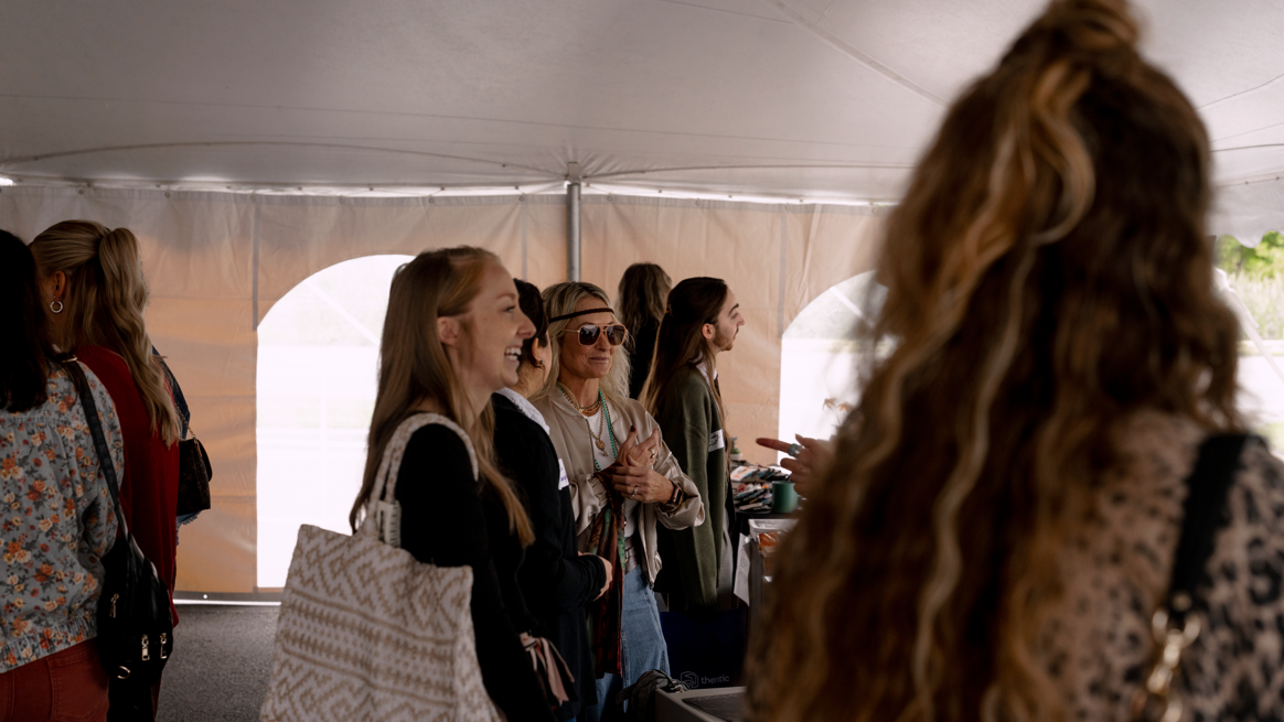 A group of women standing inside a tent, smiling and talking, with some wearing sunglasses and carrying bags.