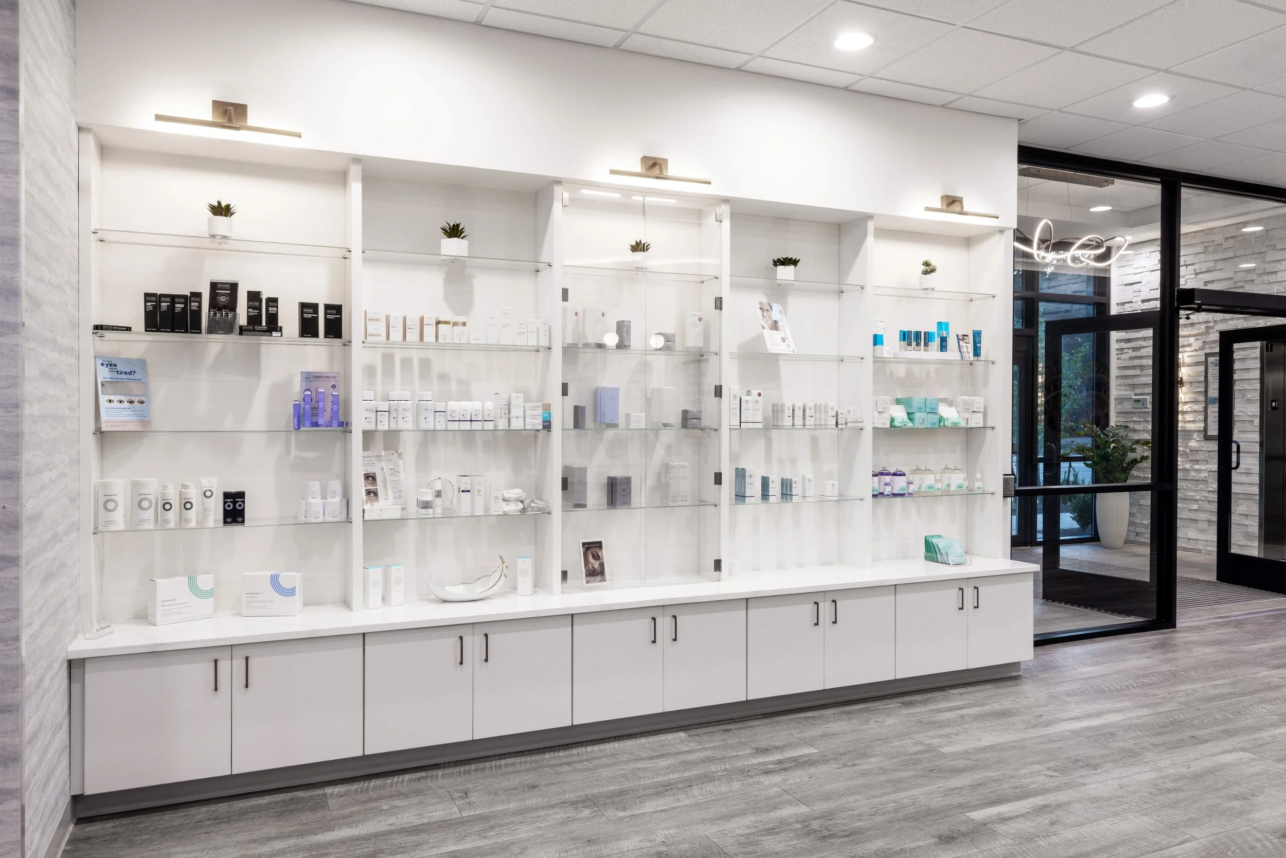Interior view of a retail display area with white shelving containing skincare and beauty products, small potted plants on top, and a nearby glass entrance door.