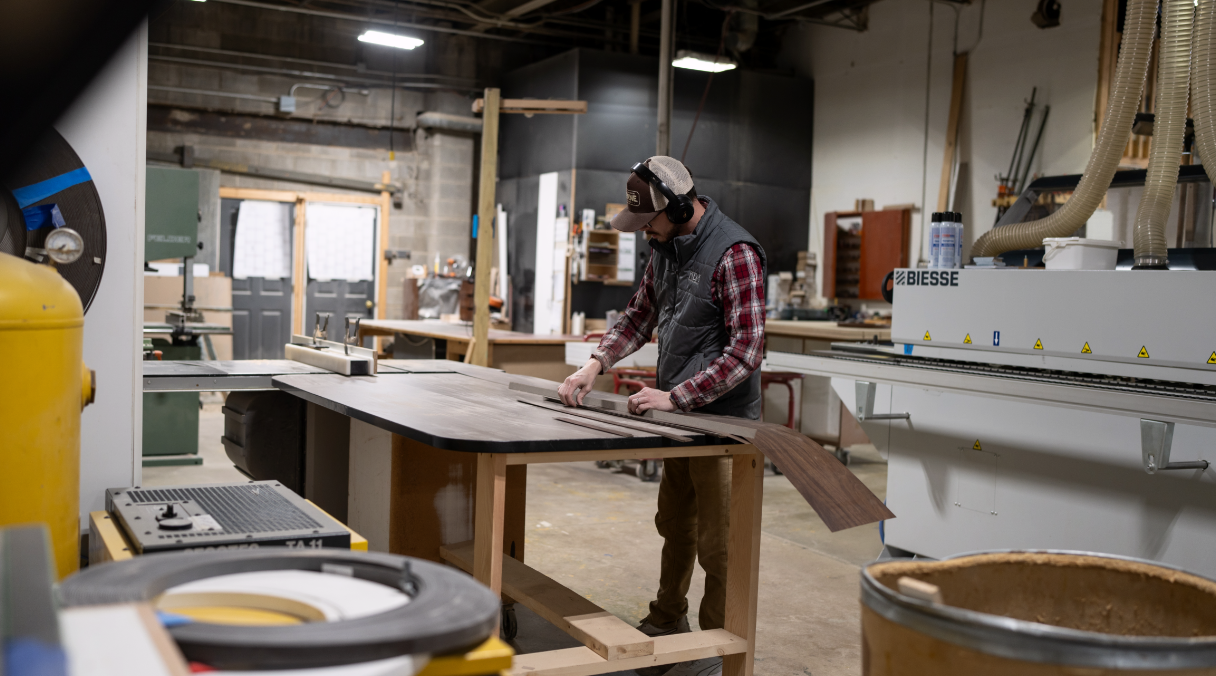 Man working on woodworking project in a workshop, wearing headphones, cap, glasses, a plaid shirt, and vest, using a ruler on a large wooden board.