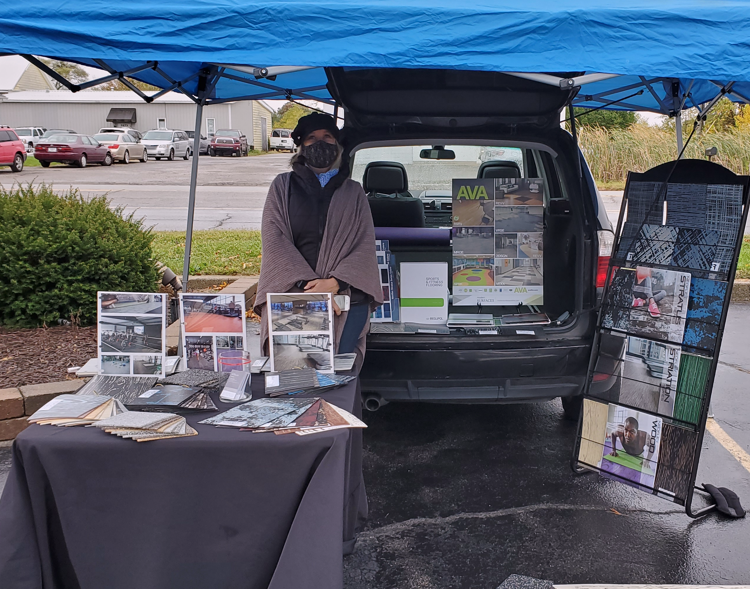 Outdoor booth with a woman wearing a mask and cape, displaying books and posters, set up in a parking lot under a blue canopy.
