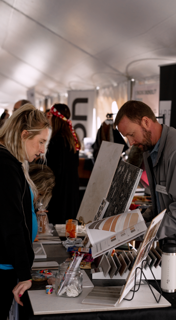 A man and a woman are looking at display samples at a trade show booth inside a tent, with other attendees in the background.