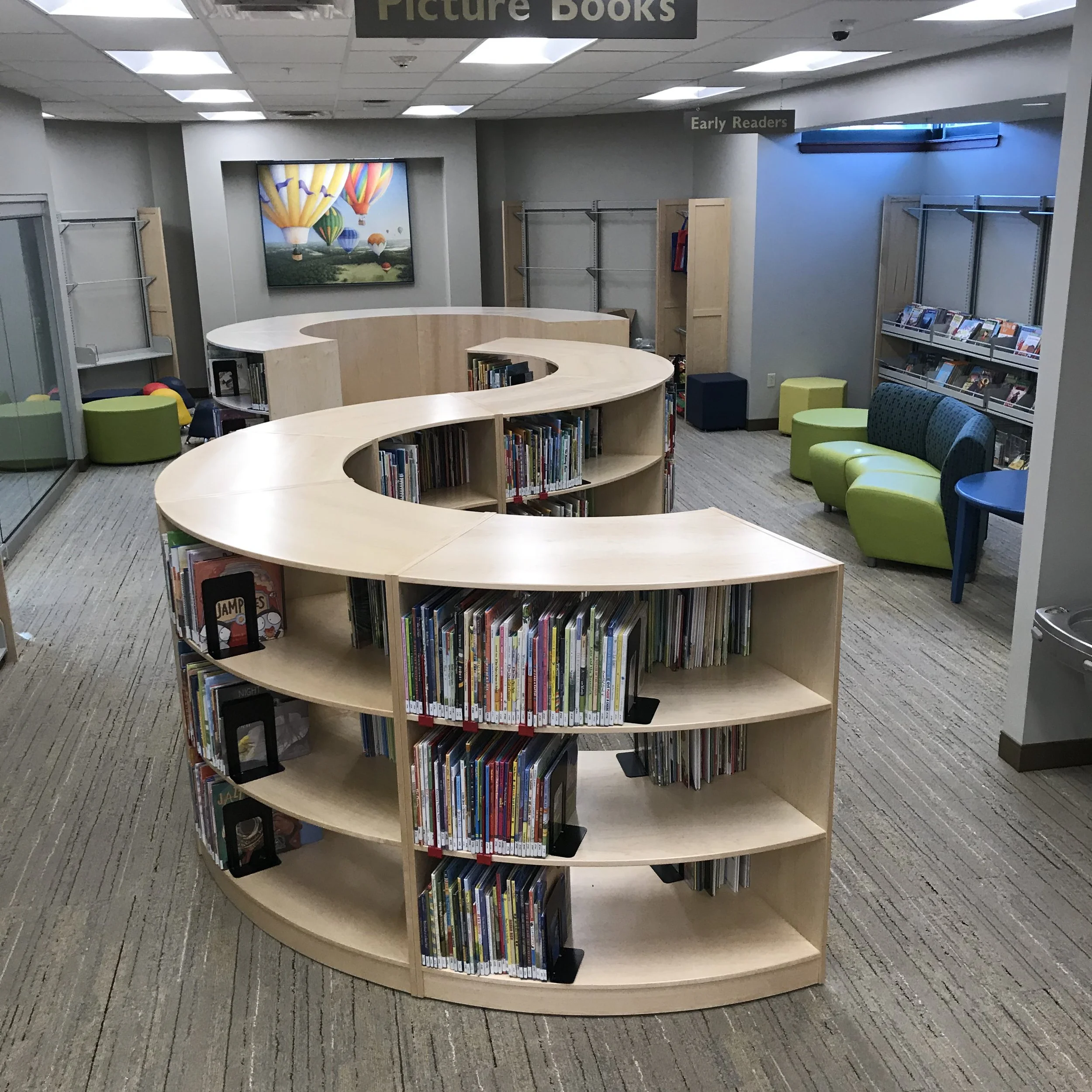 A round wooden bookshelf filled with children's books inside a library, surrounded by green and blue chairs, with signs overhead indicating sections for 'Picture Books' and 'Early Readers'.