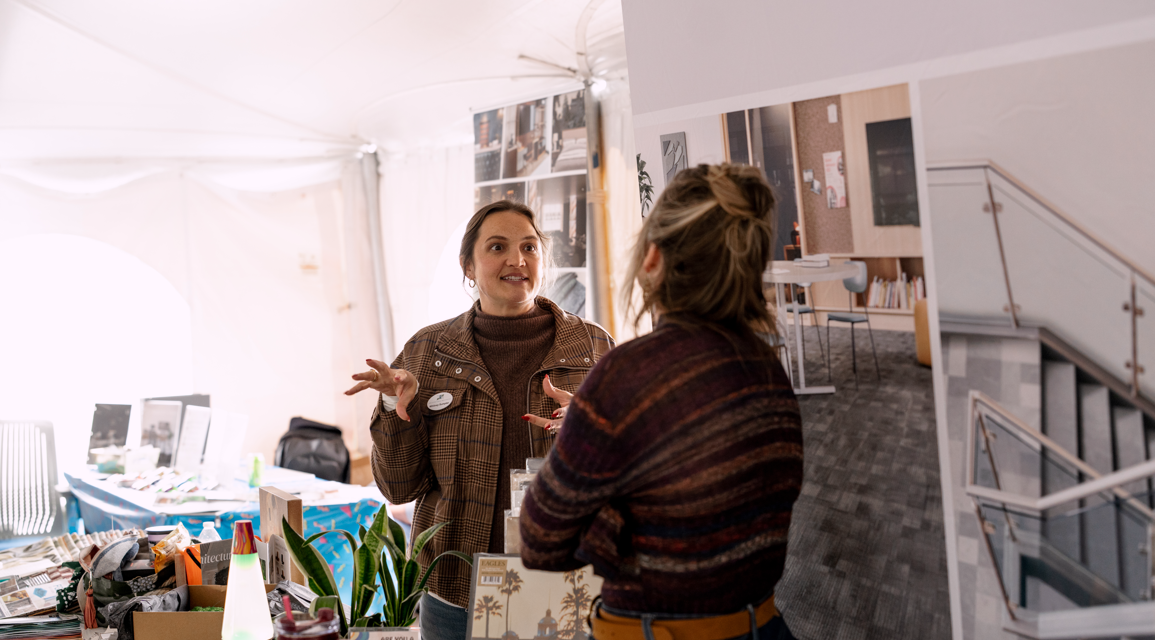 Two women conversing inside a well-lit space with tables and display items, one woman with a plaid jacket and the other with a striped sweater, near a staircase with glass railing.